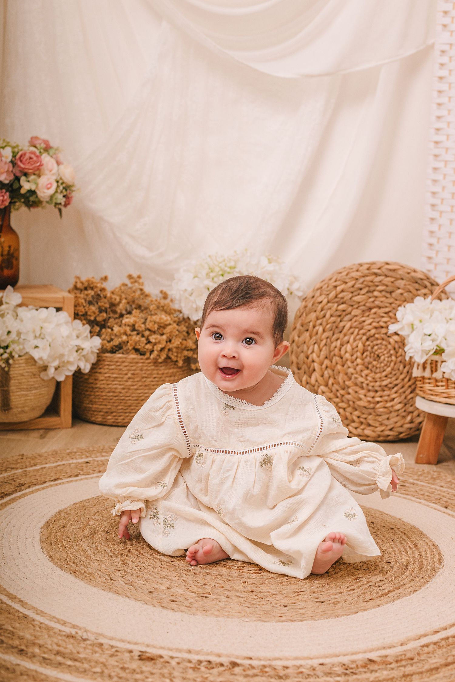 A baby in a white vintage dress sits on a round jute rug surrounded by floral decorations and woven baskets.