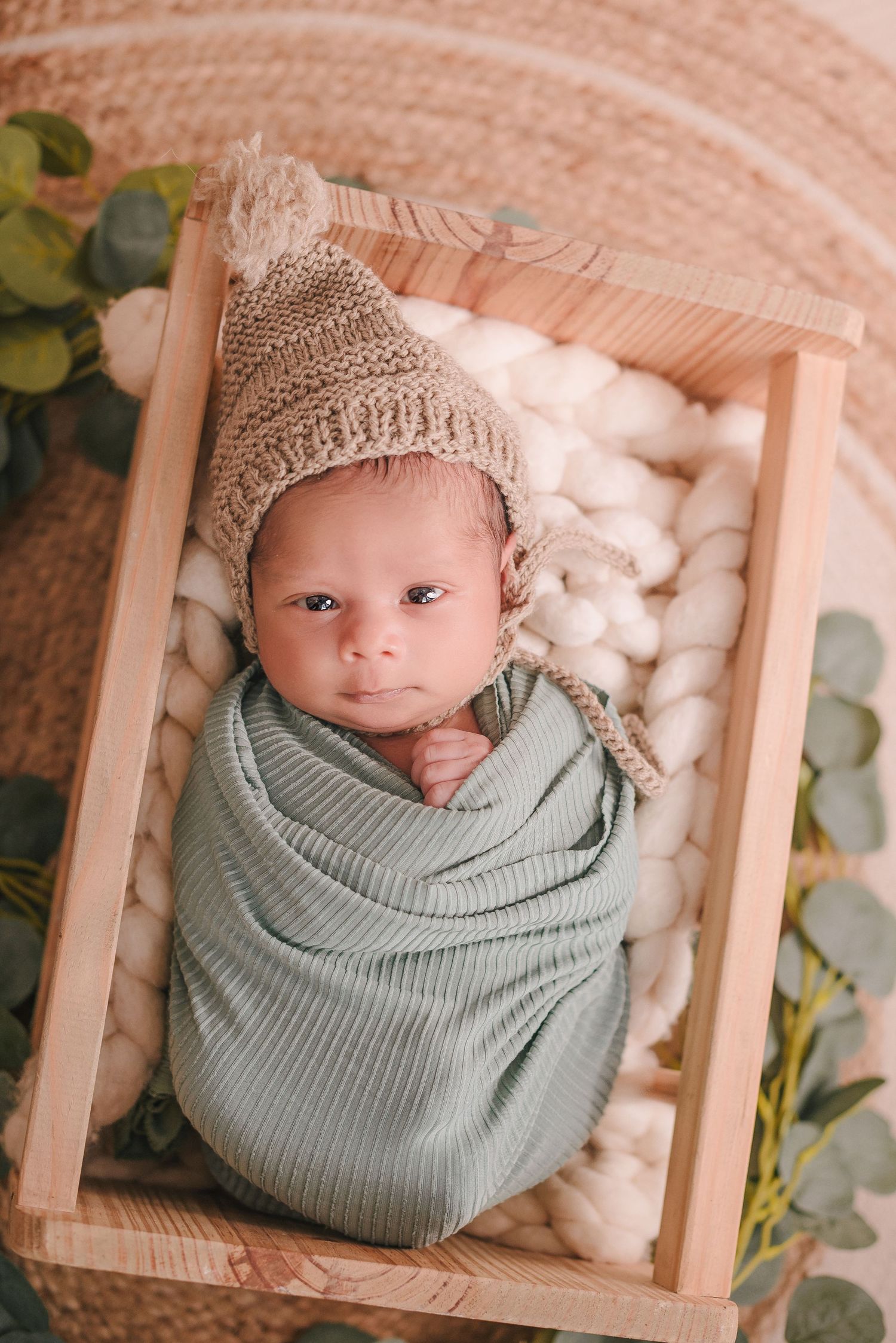 Newborn swaddled in soft sage green blanket wearing knit beanie lies in wooden crate filled with cotton.