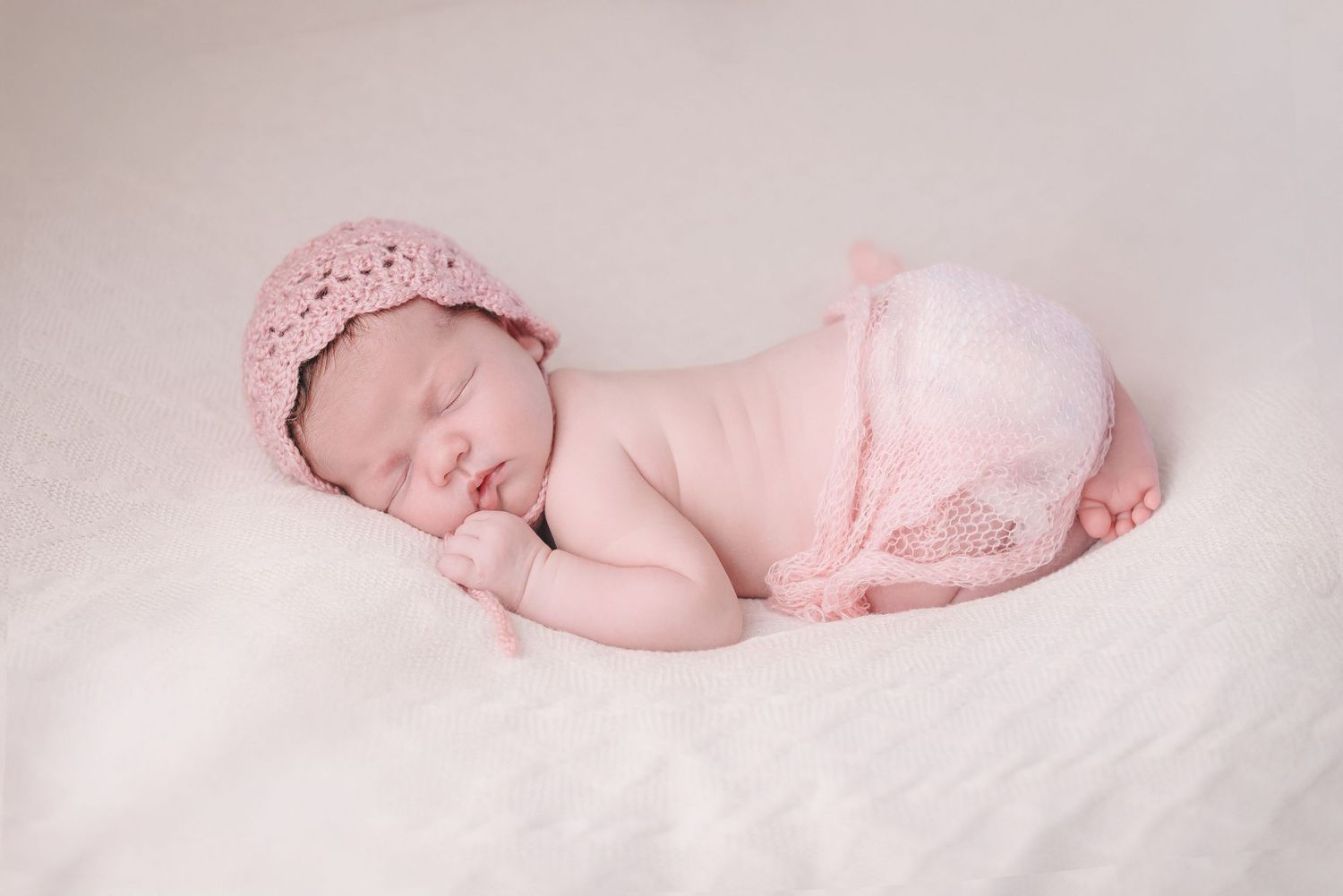 Dreamy newborn photography featuring soft pink clothing and white textured backdrop.