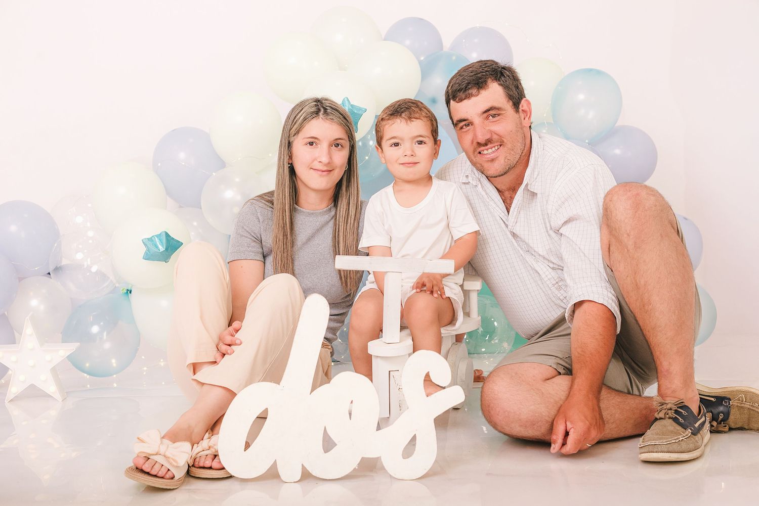 A family in white outfits poses together on a light backdrop with blue balloons and a white 'love' sign.