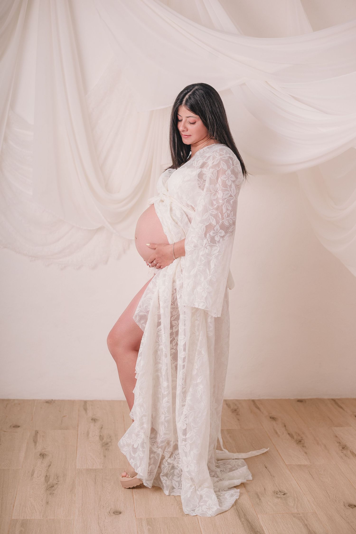 Woman in white lace robe poses elegantly during maternity session against white draped backdrop.