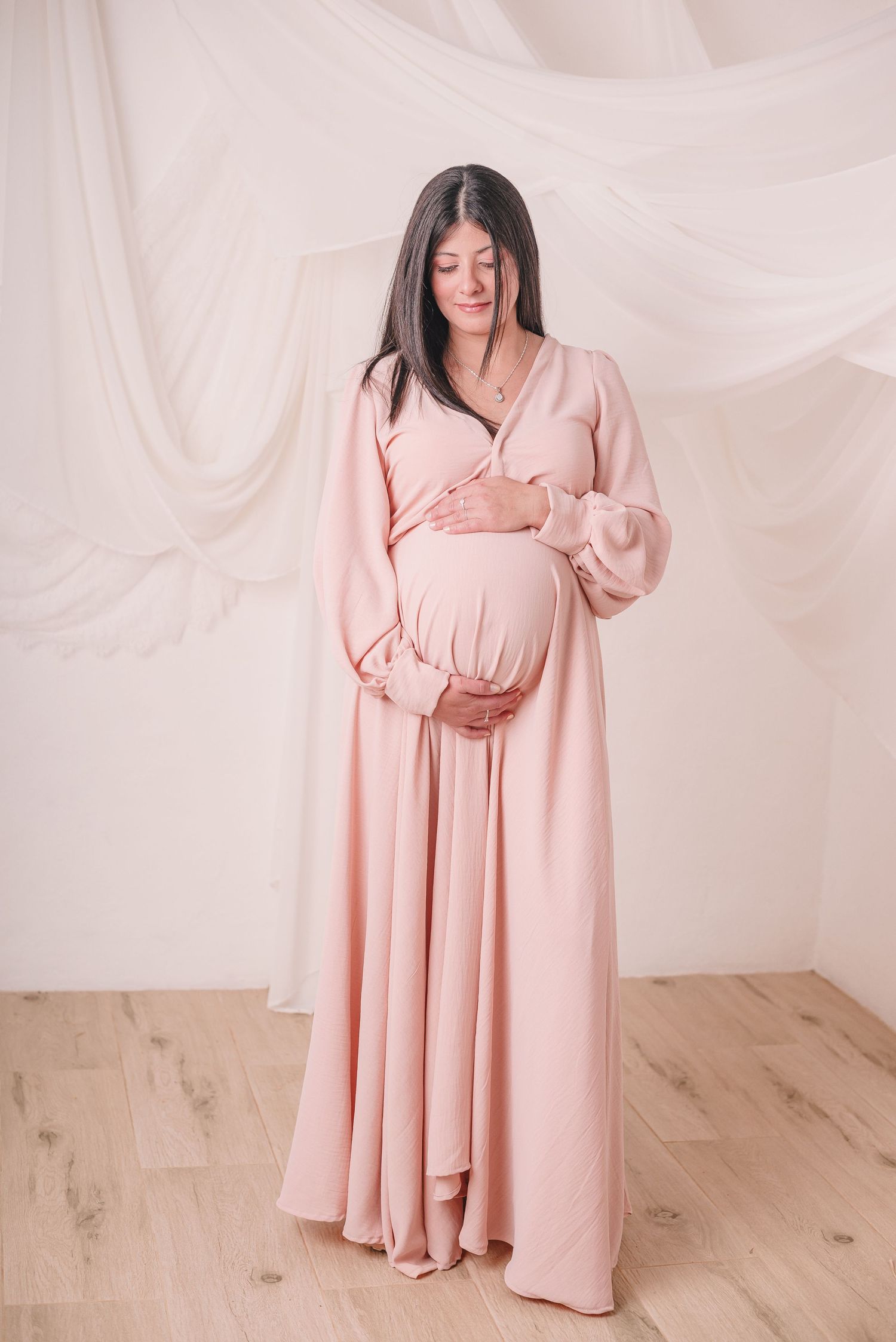 Pregnant woman in flowing pink maxi dress poses against white draping for maternity photos.