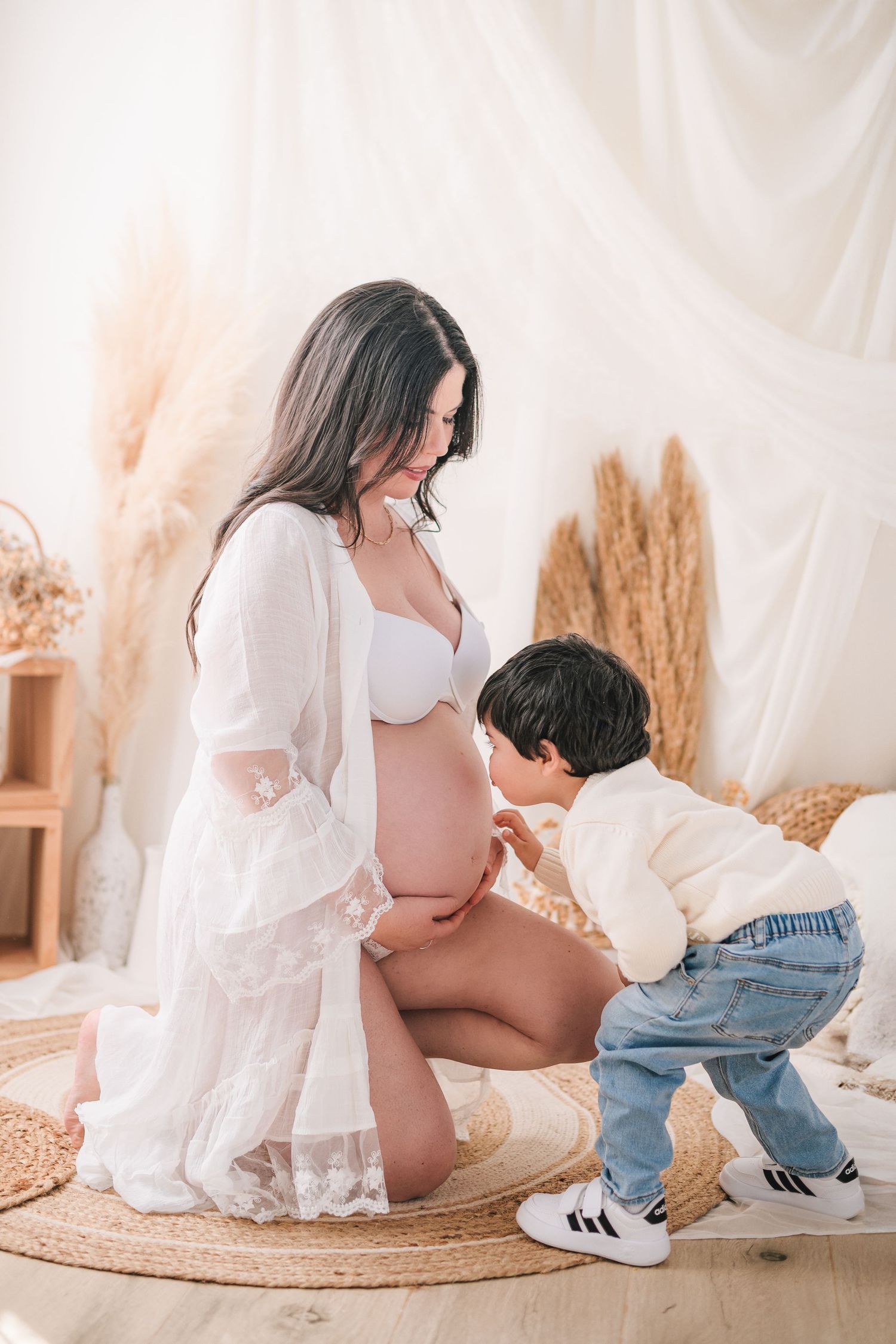 Intimate maternity photoshoot series with mother and child in white dress against neutral backdrop with dried pampas grass.