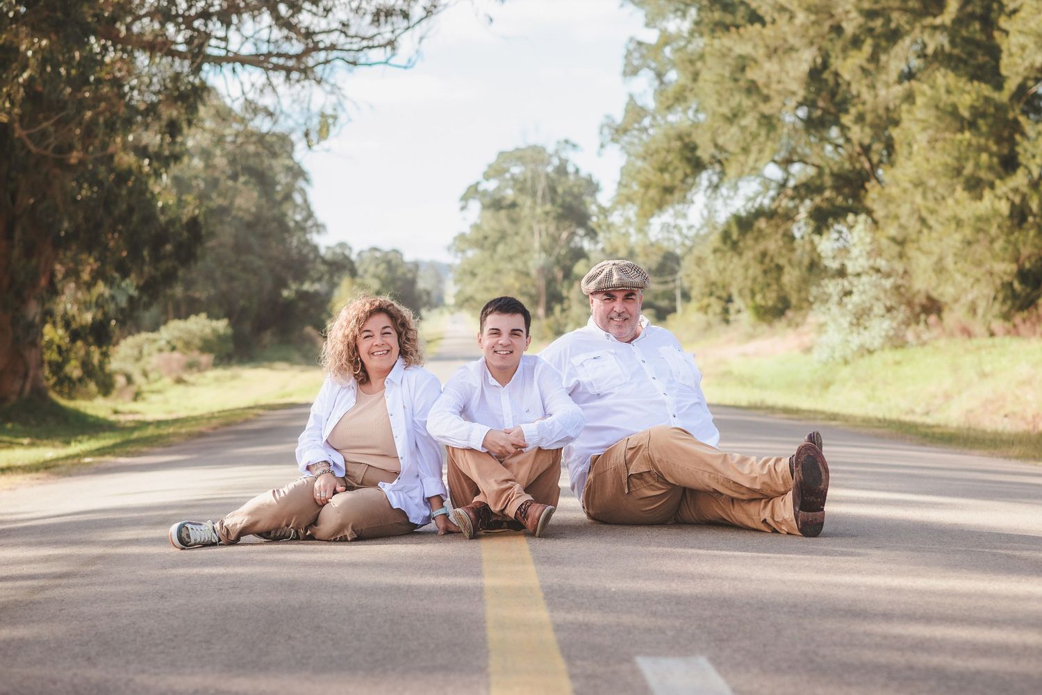 Three people in white shirts sit together on a rural road lined with trees on a sunny day.