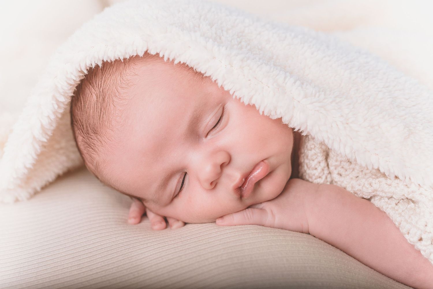 A sleeping newborn baby wrapped in white lace swaddle blanket lies peacefully on a soft textured surface.