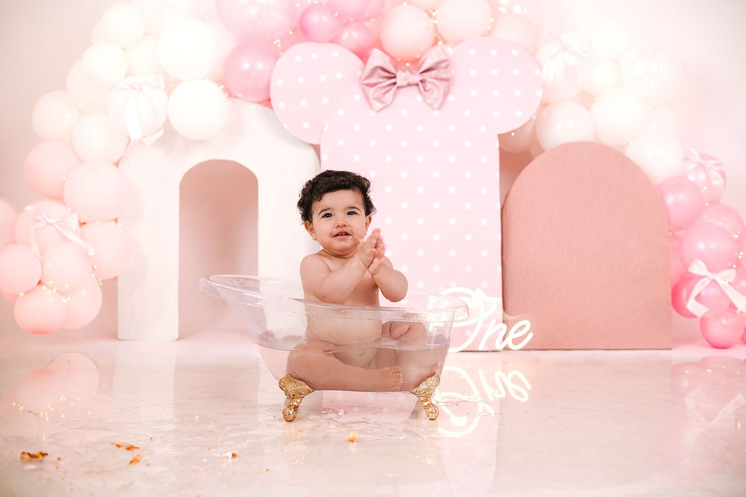 Baby girl in pink tutu sits in mini bathtub surrounded by pink balloons and number one backdrop for first birthday.