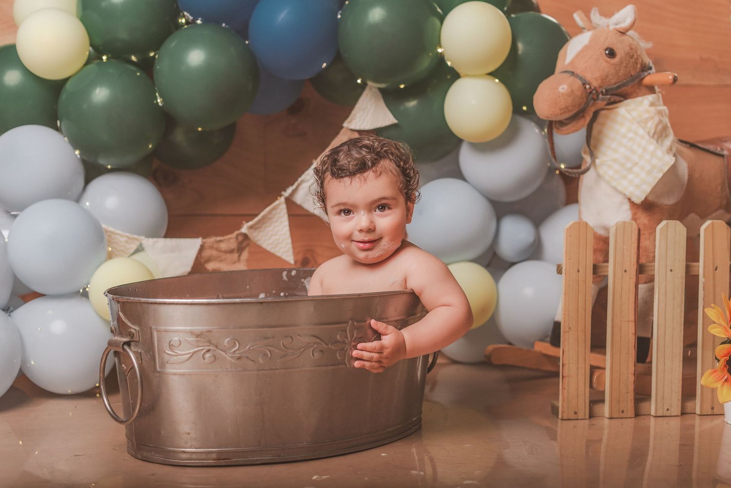 A baby sits in a metal tub surrounded by blue and green balloon decorations and rustic farmhouse decor.