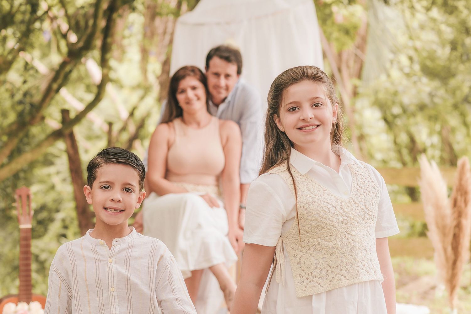 A family in light colored clothing poses together outdoors in a sun-dappled wooded setting.