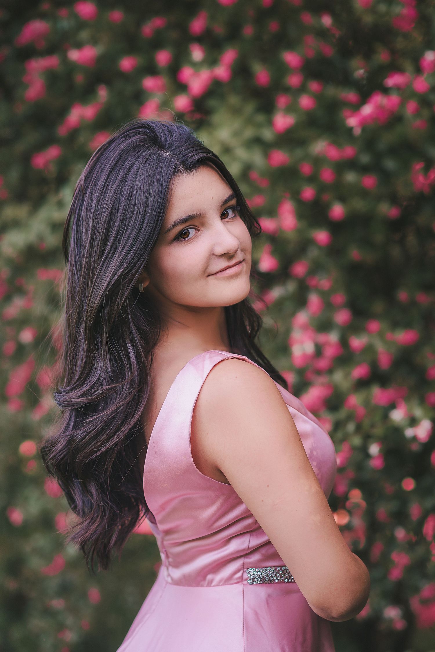 A pink formal dress stands out against a backdrop of blooming pink flowers and greenery in an outdoor portrait setting.