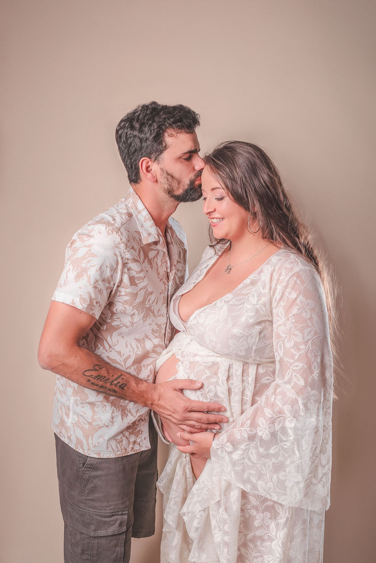 A couple in matching floral outfits share intimate moments during a maternity photoshoot against a neutral backdrop.