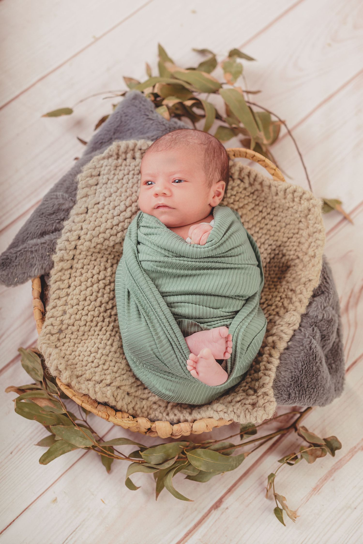 A newborn baby swaddled in sage green wrap lies on textured beige blanket surrounded by scattered eucalyptus leaves.