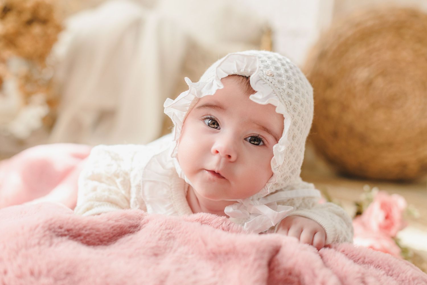 Baby wearing white lace bonnet lies on soft pink blanket in gentle natural lighting.