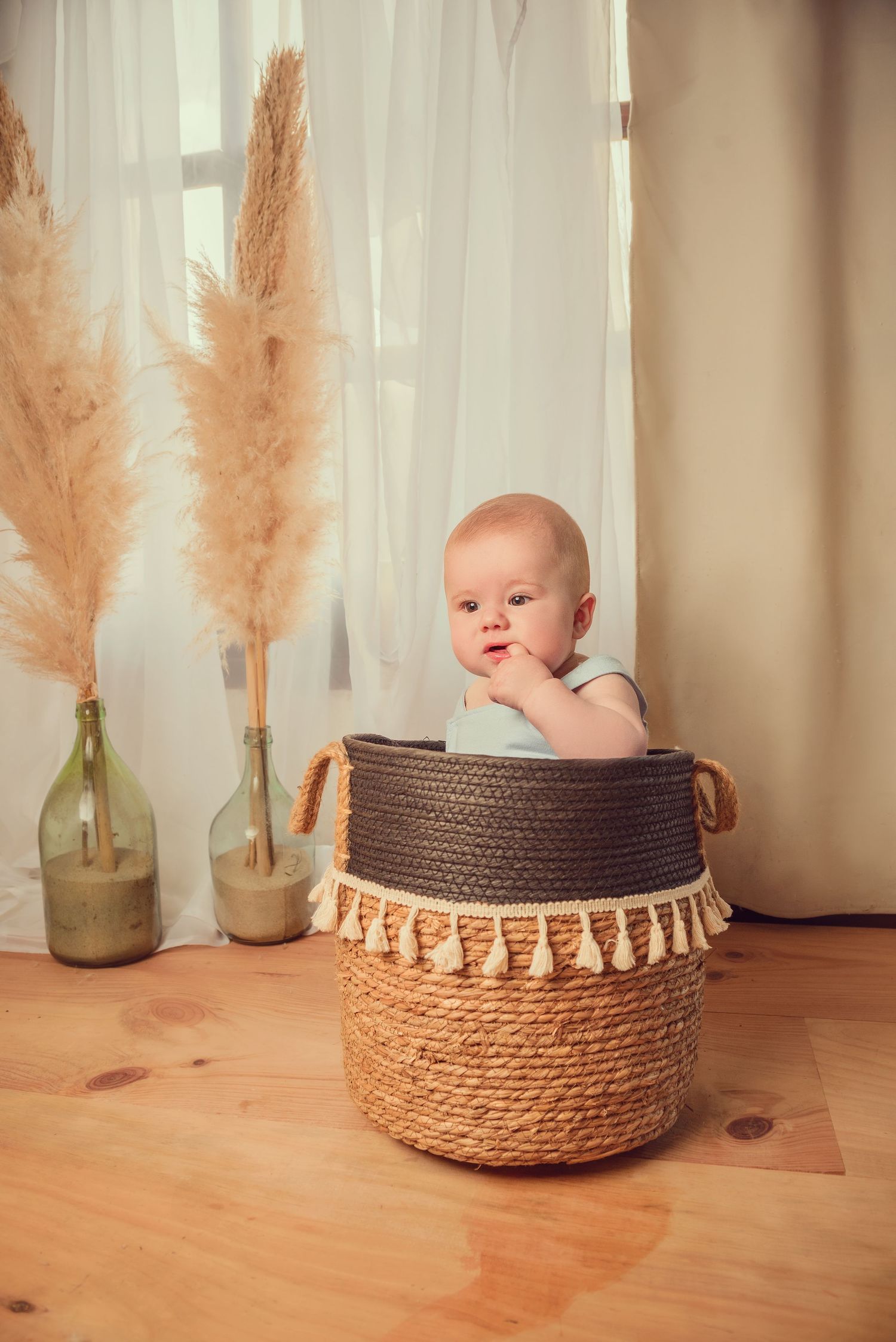 Baby sits in woven basket with pampas grass decor and soft window light creating cozy nursery scene.