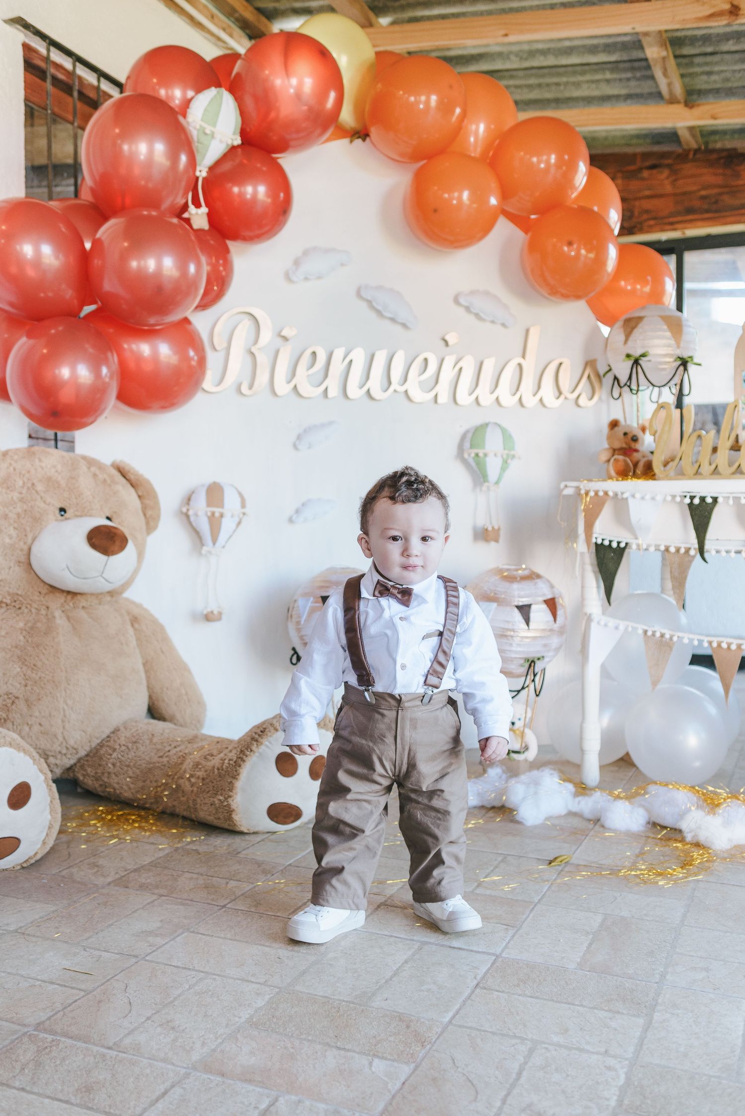 Baby in beige outfit with suspenders playing under orange and white balloon arch decoration at birthday party.
