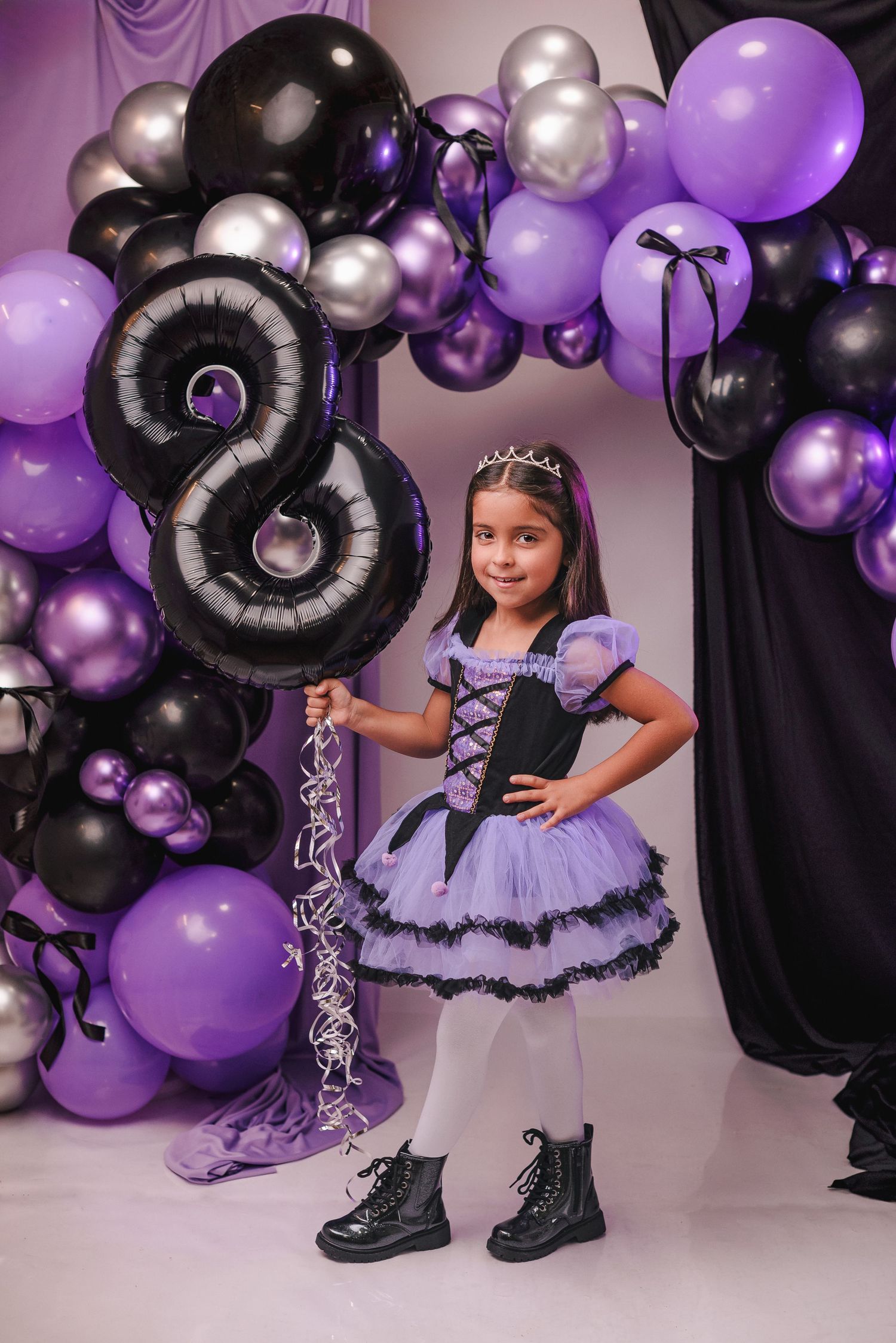 A purple and black themed birthday photoshoot setup with balloons, tulle dress, and combat boots for a young child.