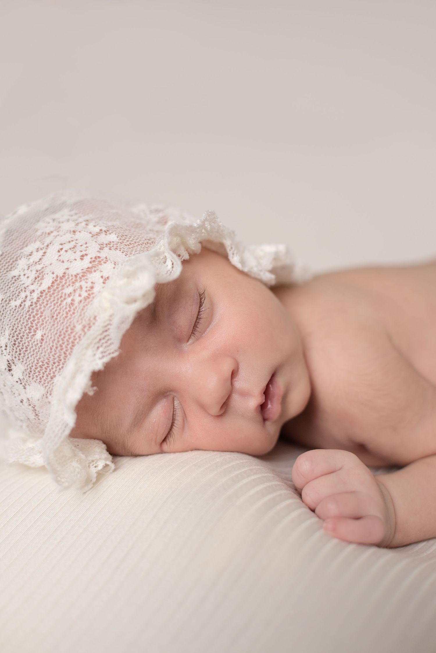 A sleeping baby wearing delicate white lace lies peacefully on soft white bedding.