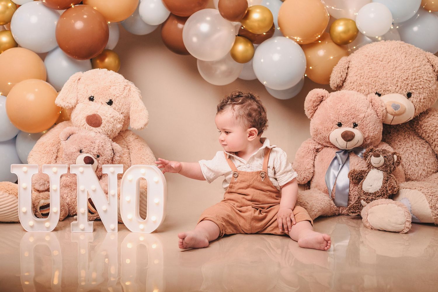 A baby sits on the floor surrounded by large teddy bears and balloon decorations with 'UNO' letters for first birthday.