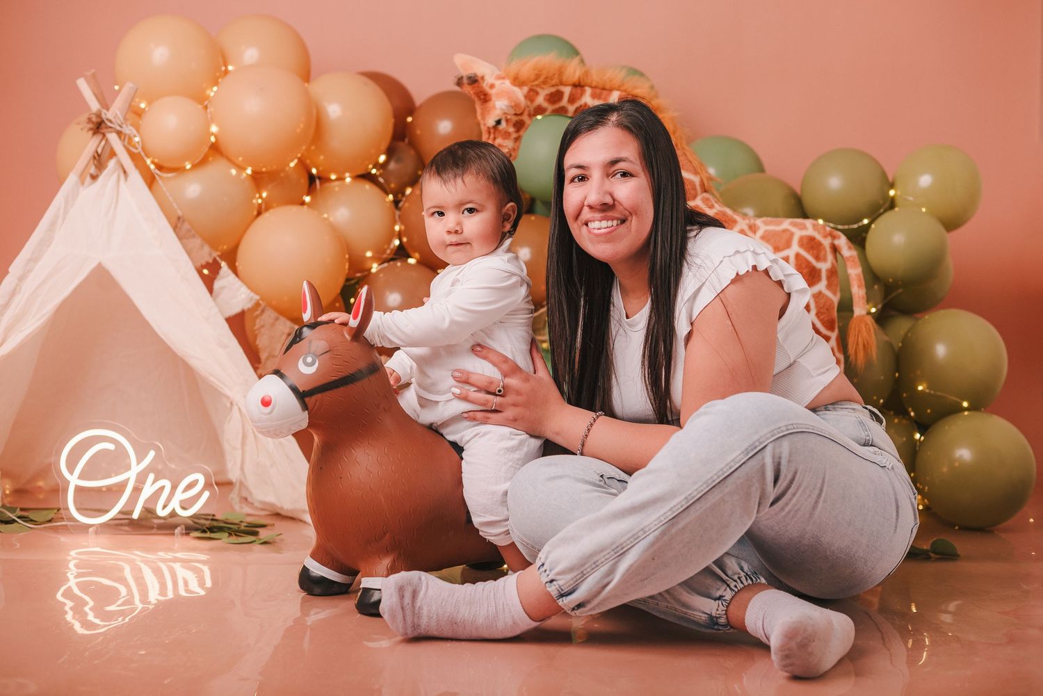 Baby in white dress poses with balloons for first birthday cake smash photo session against pink backdrop.