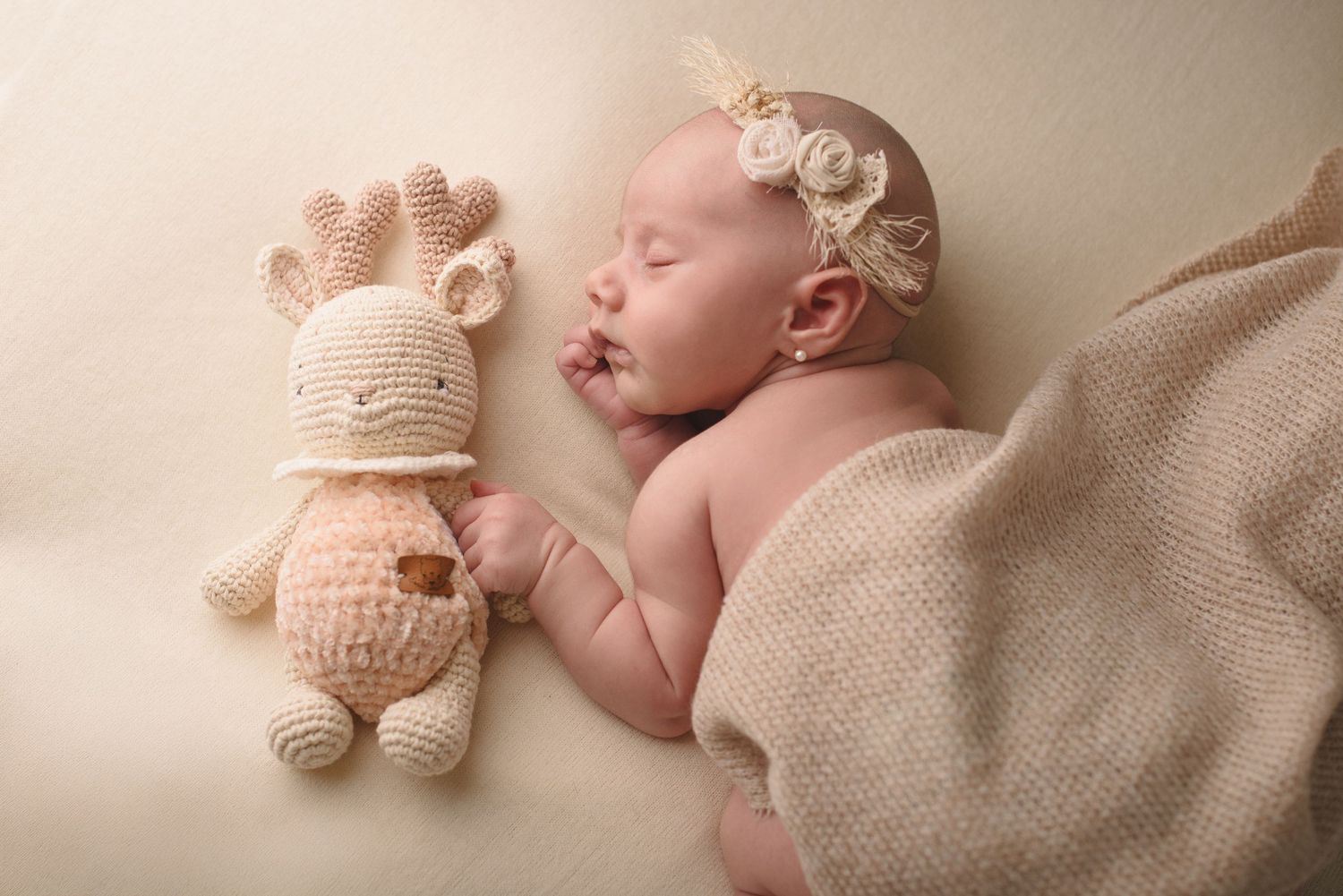 A sleeping baby in soft beige tones wearing a delicate floral headband lies peacefully next to a plush teddy bear.
