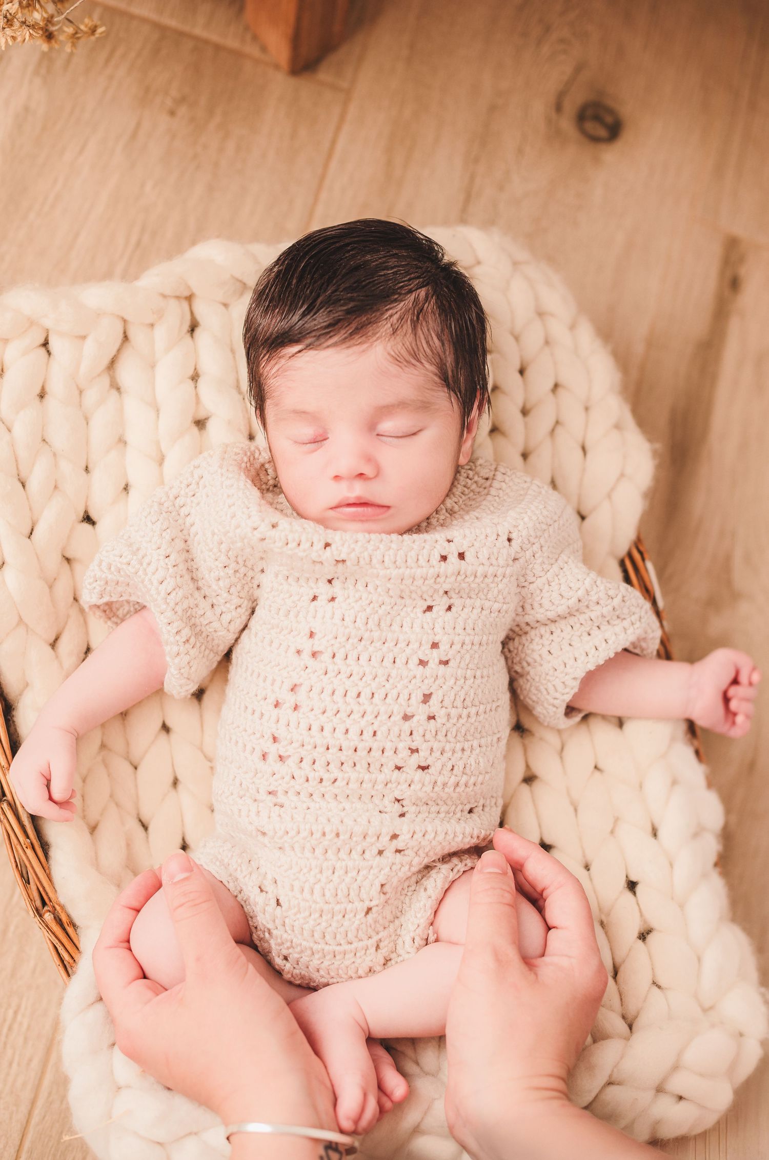 Newborn baby wearing cream knit romper sleeping peacefully on chunky knit blanket in warm natural lighting.