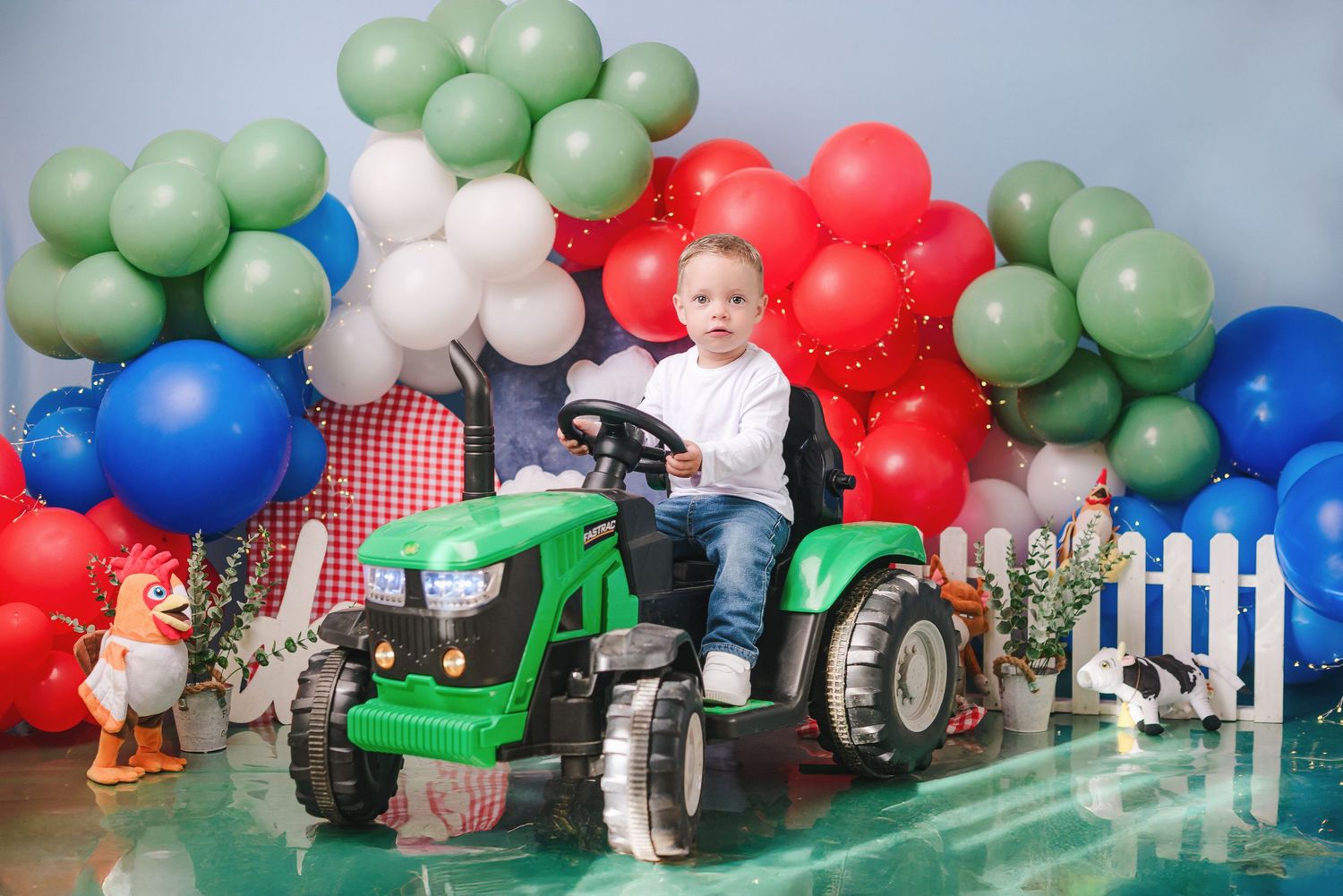 A festive balloon arch display with green, red, blue and white balloons arranged around a tractor and picket fence backdrop.