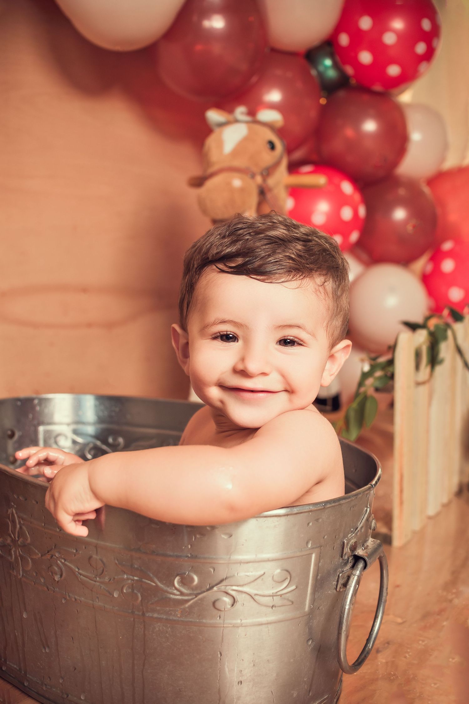 Festive holiday photo setup with red and polka dot balloons, teddy bear, and metal bucket against beige backdrop.