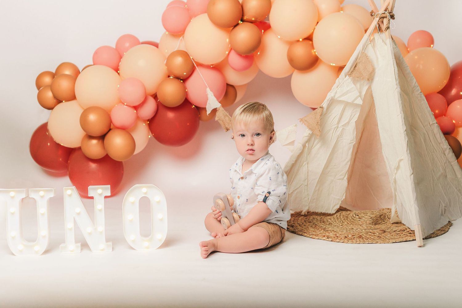 First birthday setup with teepee tent, peach and coral balloon garland, and 'UNO' text display on white backdrop.