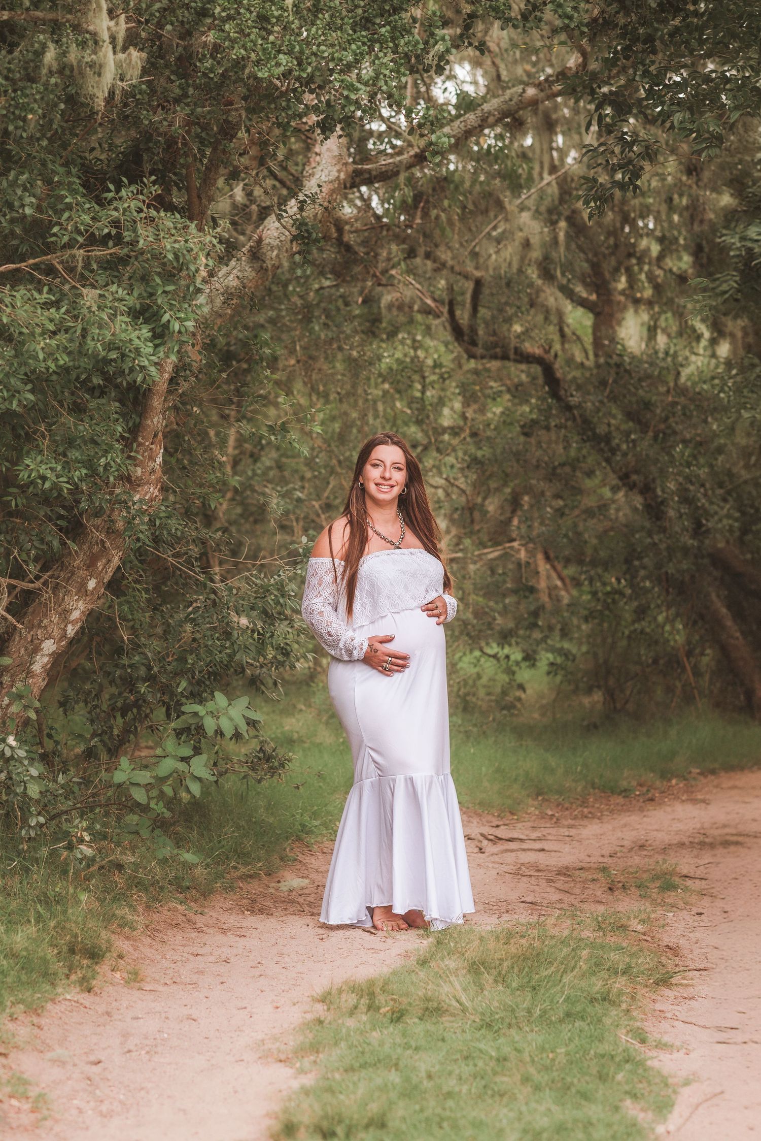 Maternity photo of expectant mother in white dress on forest path.