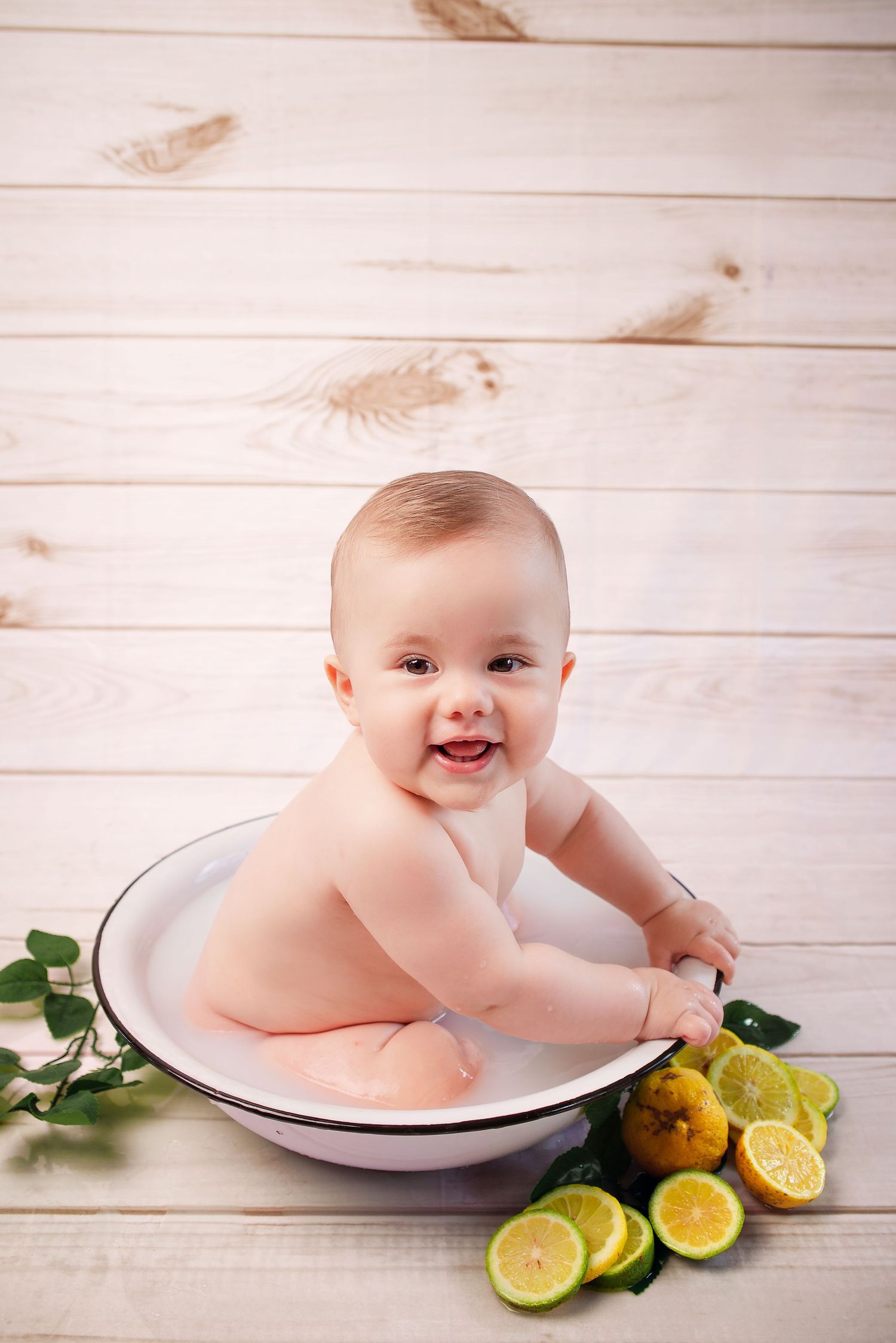 Baby sitting in white enamel basin with fresh citrus fruits on wooden plank background.