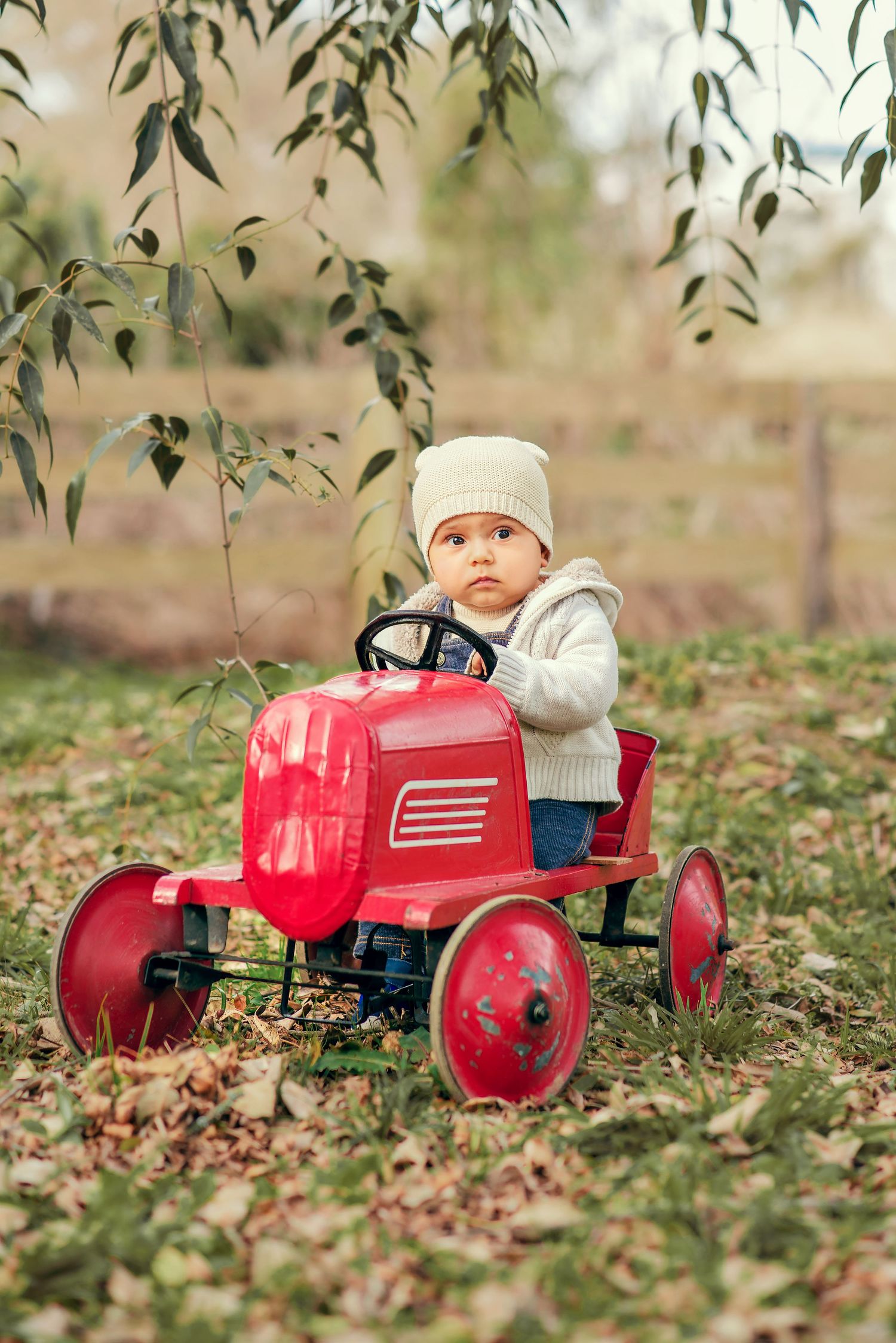 A child in winter clothing sits on a vintage red toy pedal tractor in a yard covered with fallen autumn leaves.