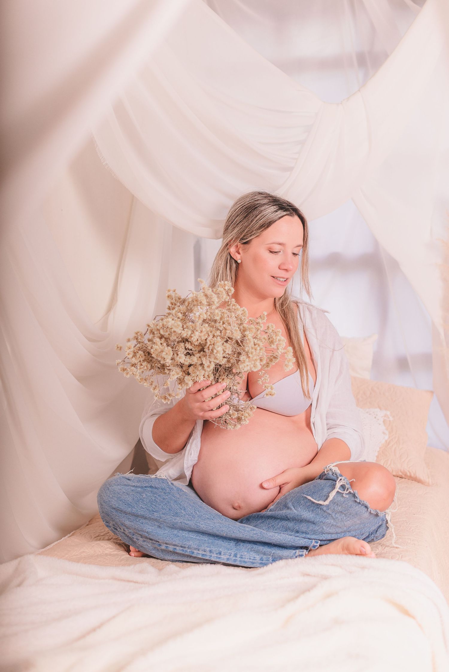 Pregnant woman in denim and cardigan holds dried flowers during indoor maternity photoshoot.