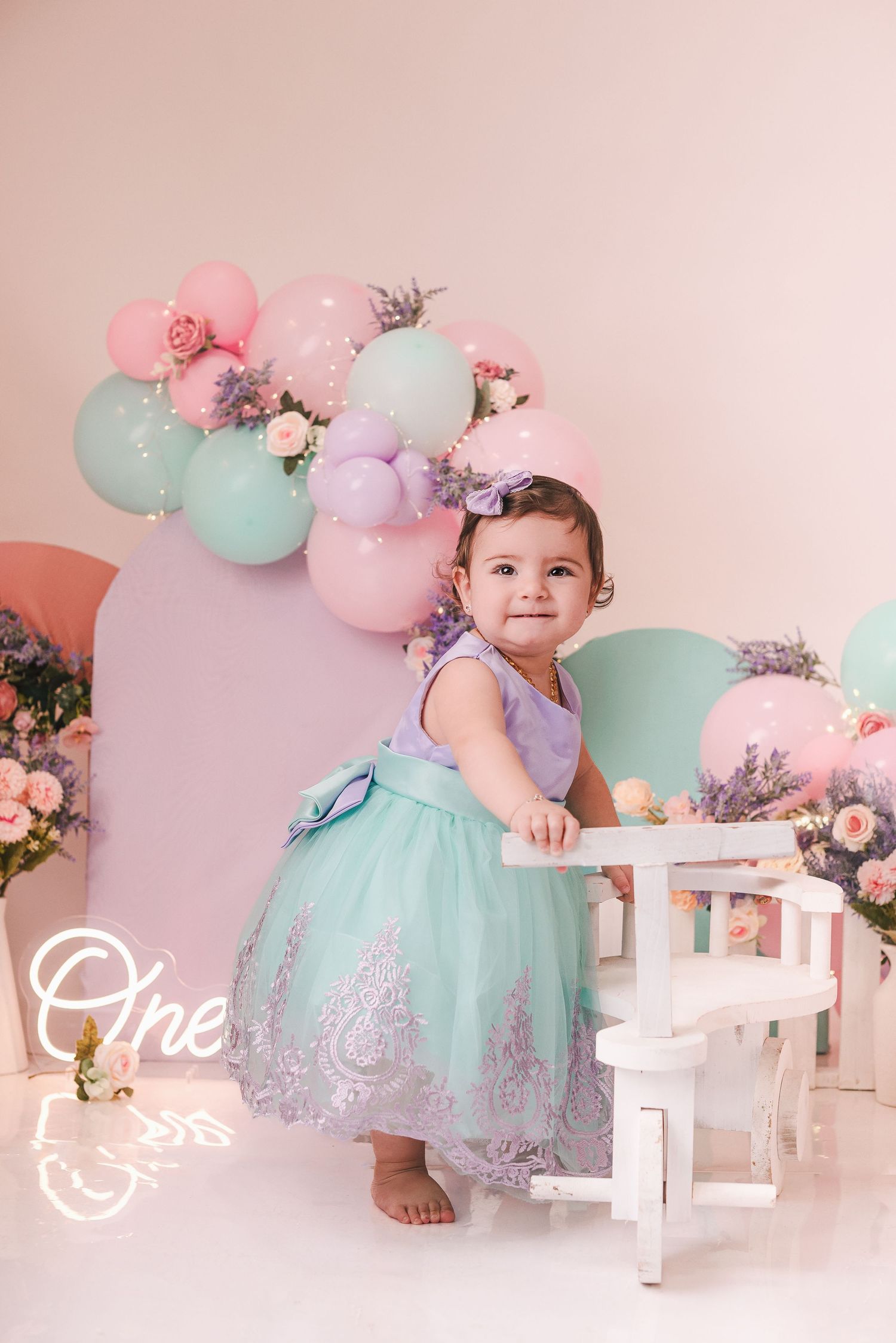 A toddler in a mint tulle dress poses for birthday photos with pastel balloons and floral decorations.