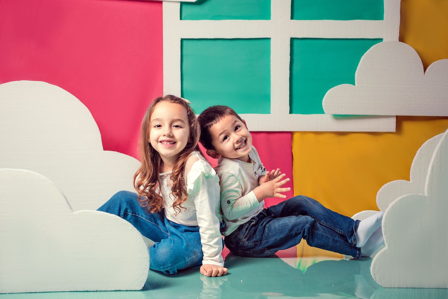 Two happy children sitting back-to-back in a colorful playroom with cloud decorations.