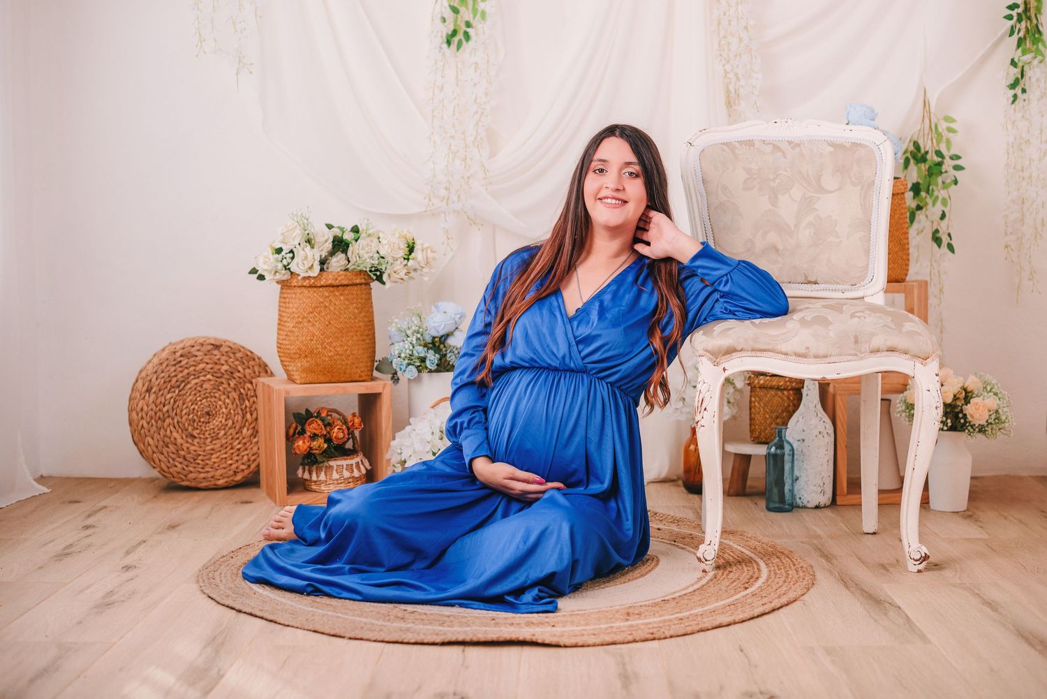 Woman in royal blue dress poses elegantly in bright studio setting with natural decor and round jute rug.