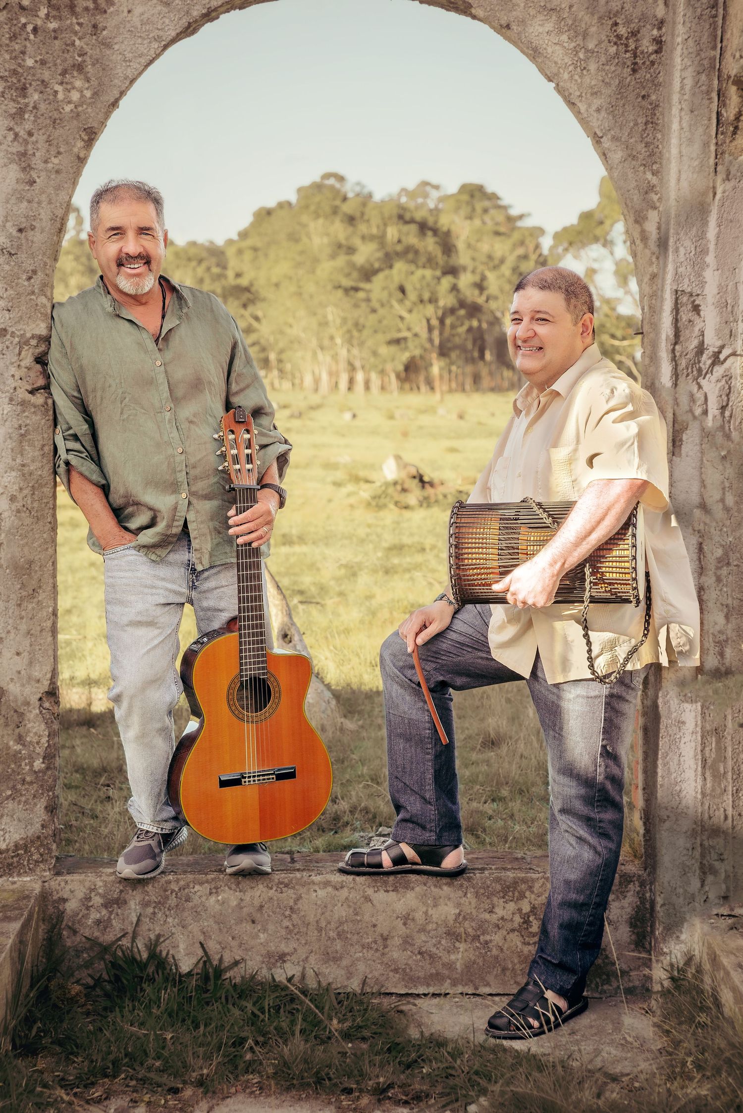 Two musicians pose with their instruments - a guitar and accordion - under a stone archway in a pastoral setting.