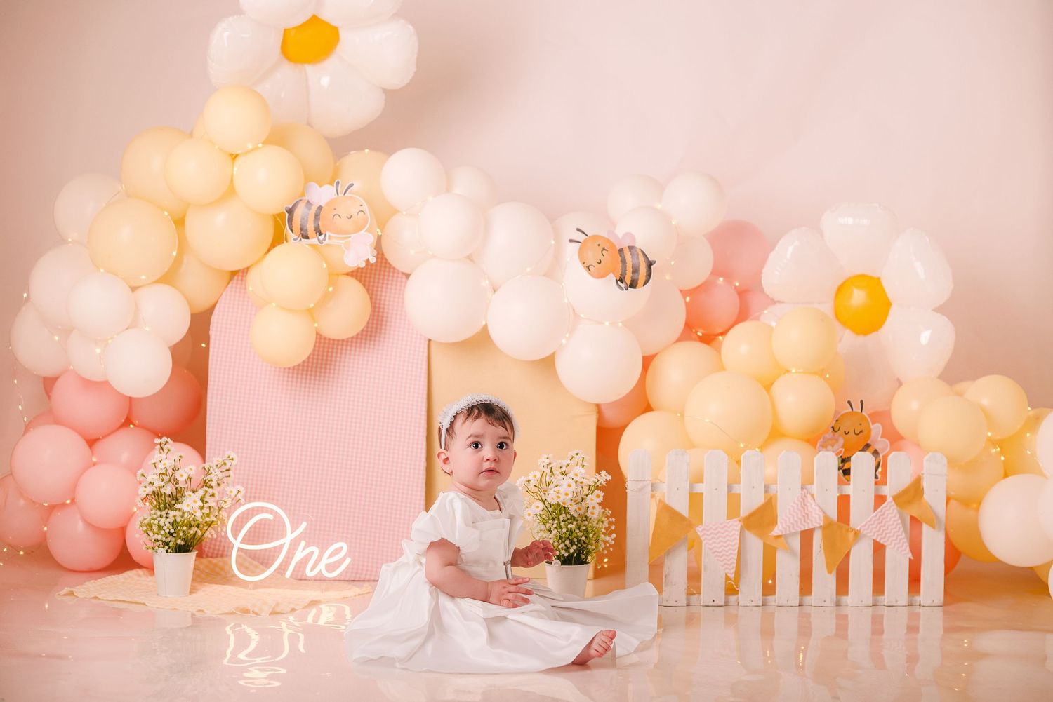 Baby's first birthday party setup with pastel balloon arch and white picket fence decorations.