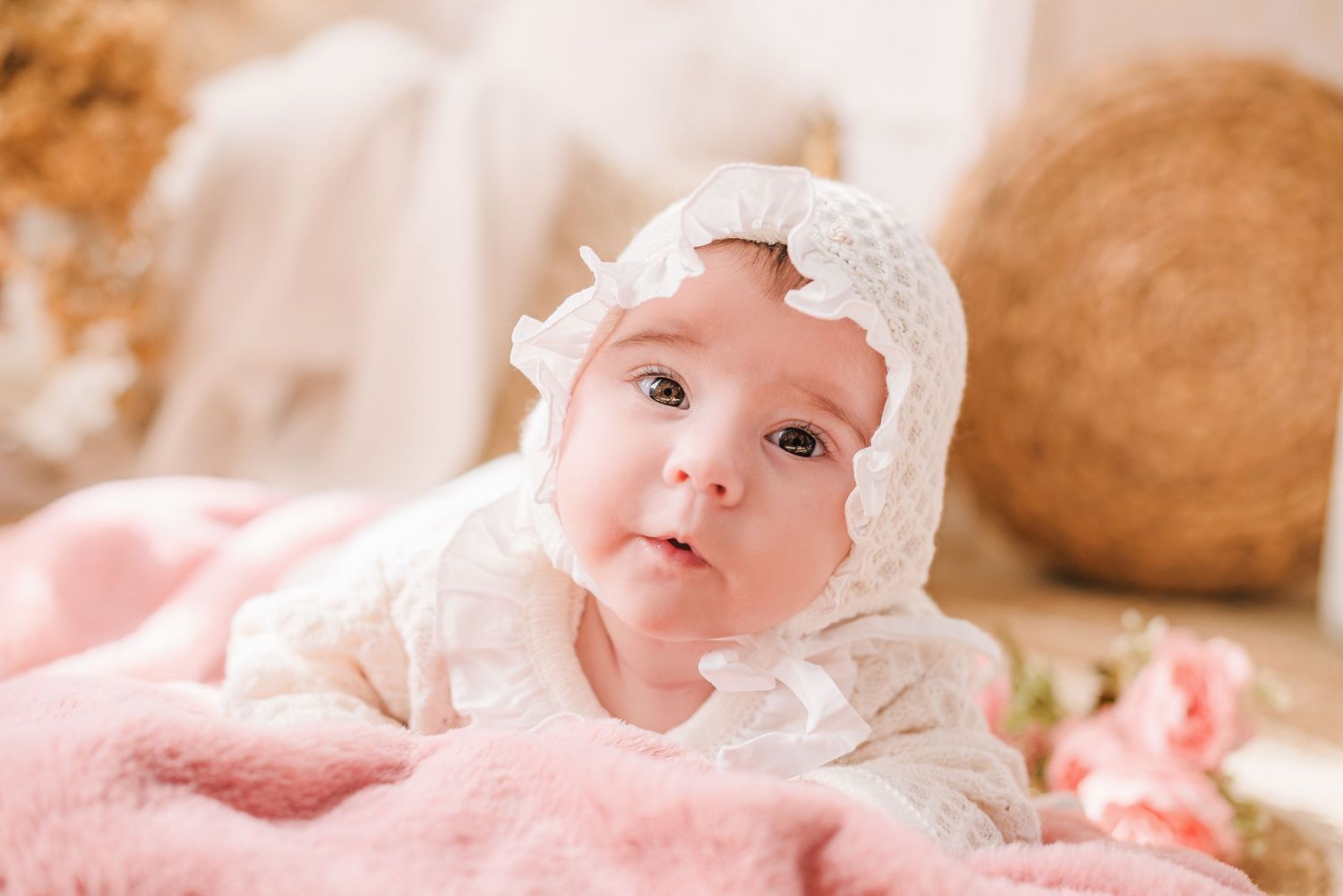 Baby wearing delicate white lace bonnet lying on pink blanket.