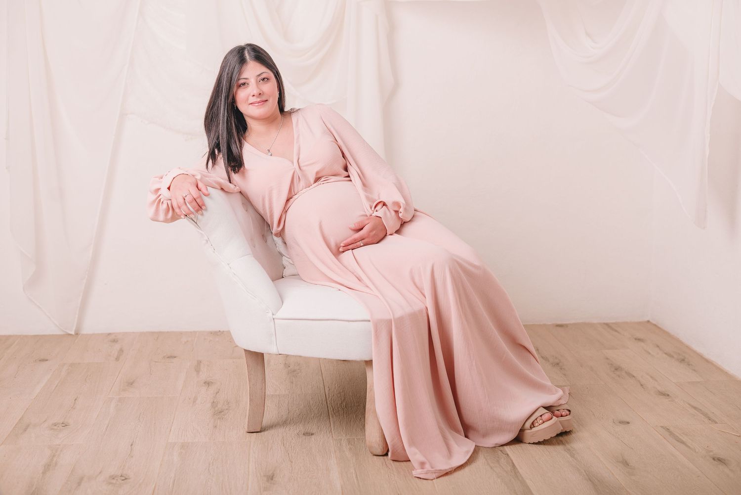 Woman in pink silk dress poses gracefully in maternity photo shoot with white fabric backdrop and wooden floor.