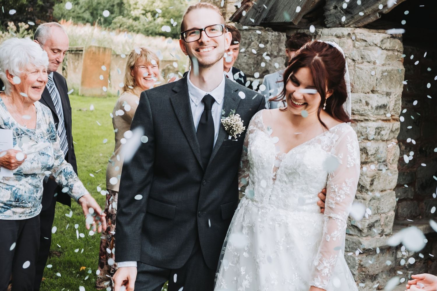 Wedding guests throw confetti over a bride and groom in formal attire outside a stone church.