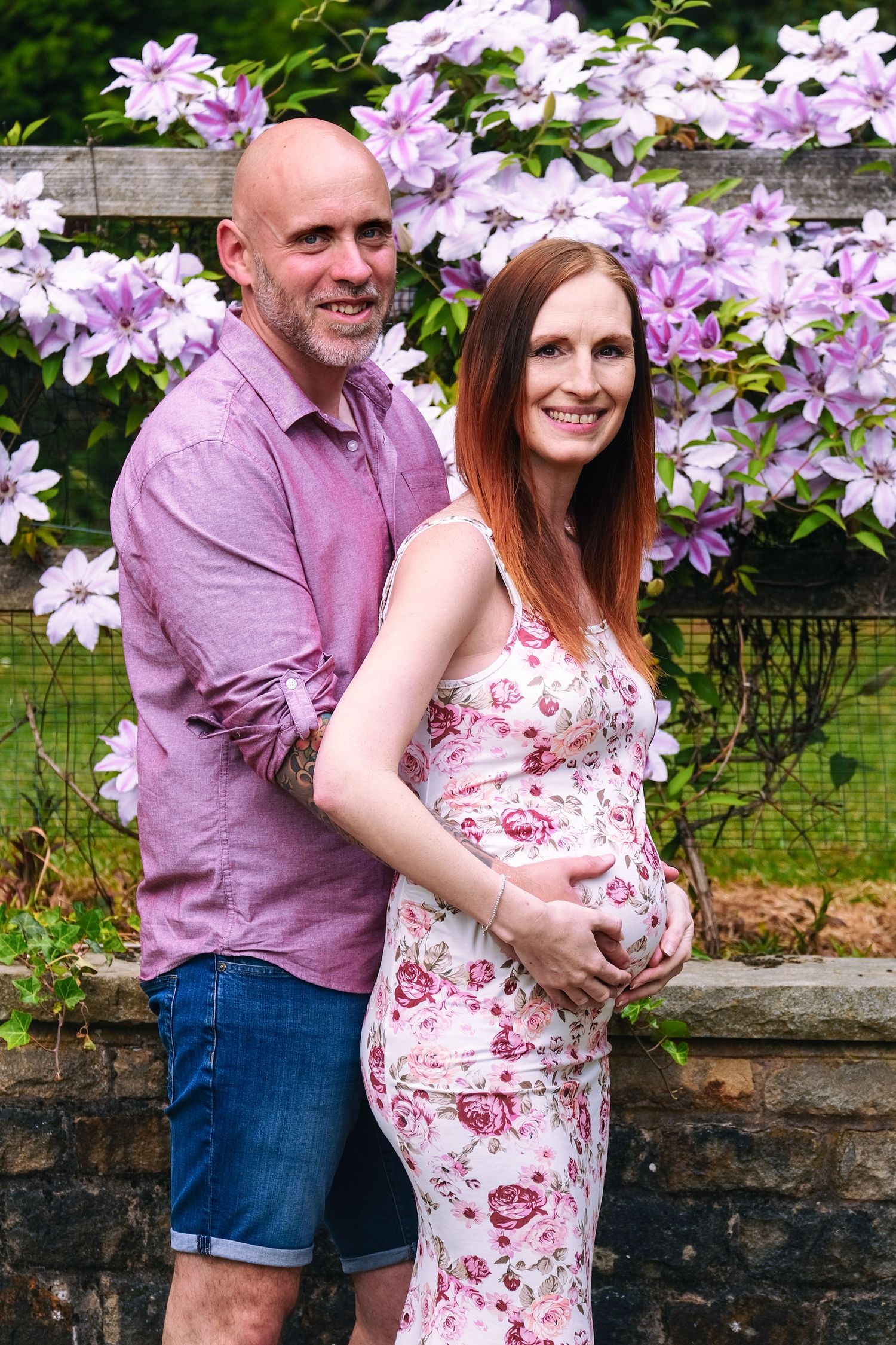 Couple embracing in front of purple clematis flowers during a maternity photoshoot.