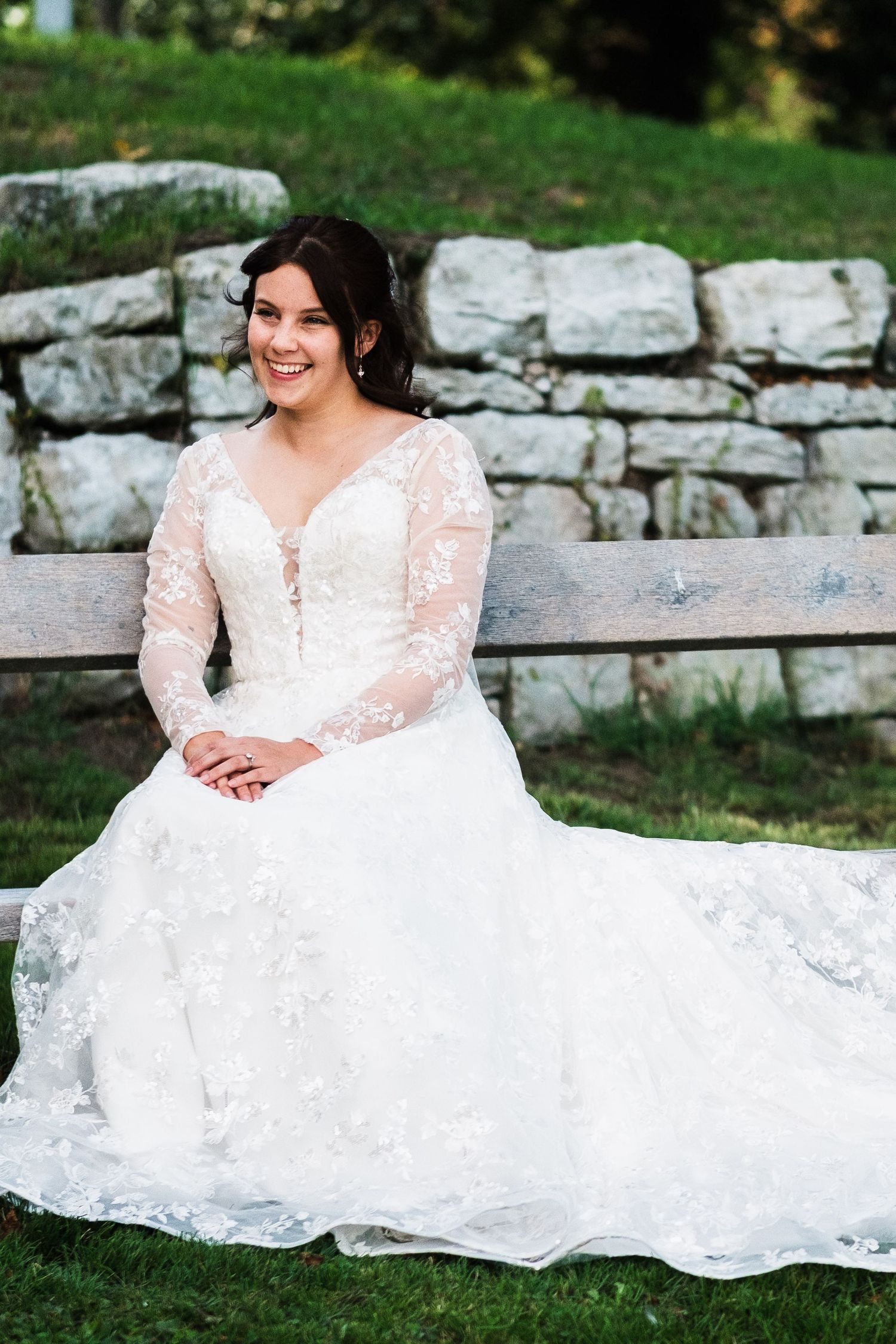 A bride in a flowing white lace wedding gown poses gracefully on stone steps in an outdoor garden setting.