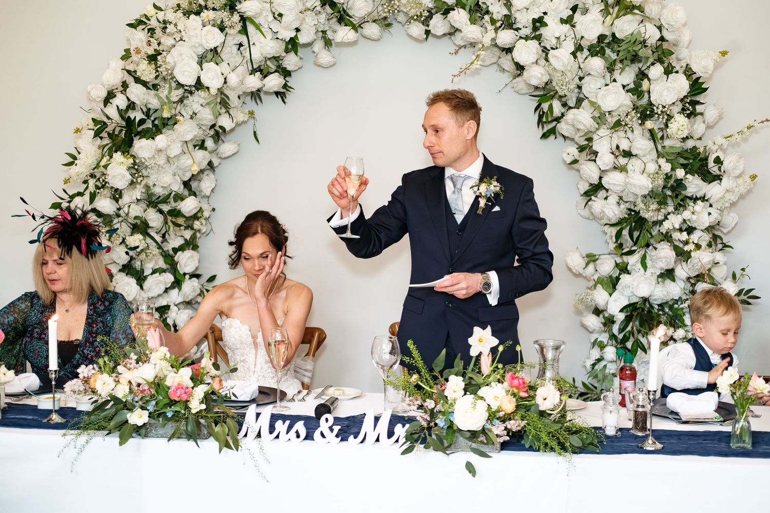 Groom raises a champagne glass during wedding speech at floral arch decorated head table with Mr & Mrs sign.