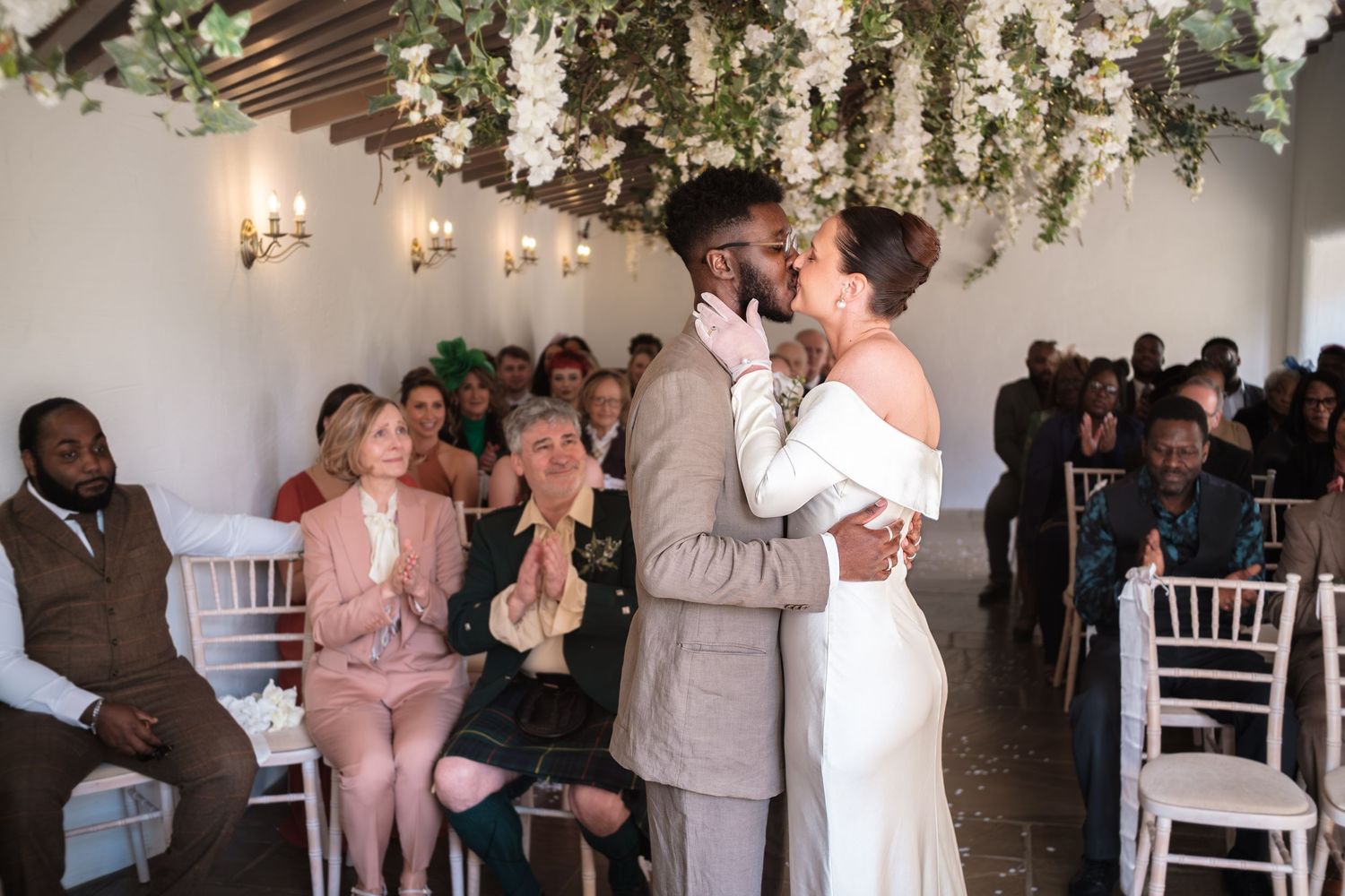 Bride and groom share first kiss at wedding altar while guests applaud in elegant white venue with floral ceiling  at dunston hall estate
