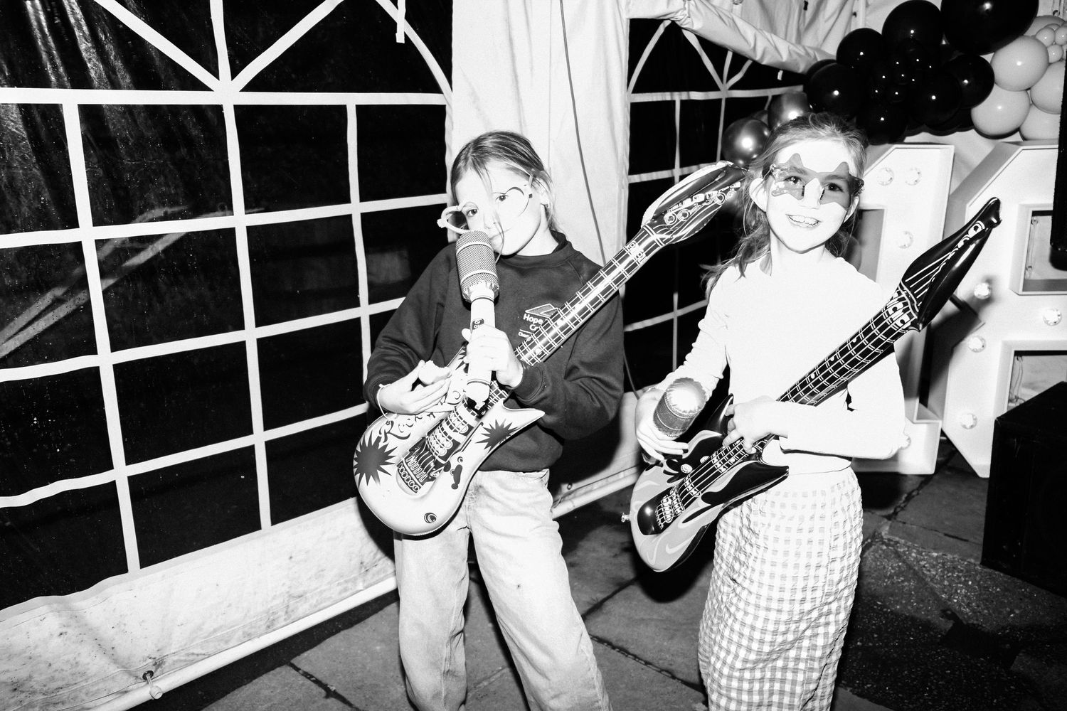 Two guitarists performing together in black and white at an indoor venue with geometric window patterns behind them.