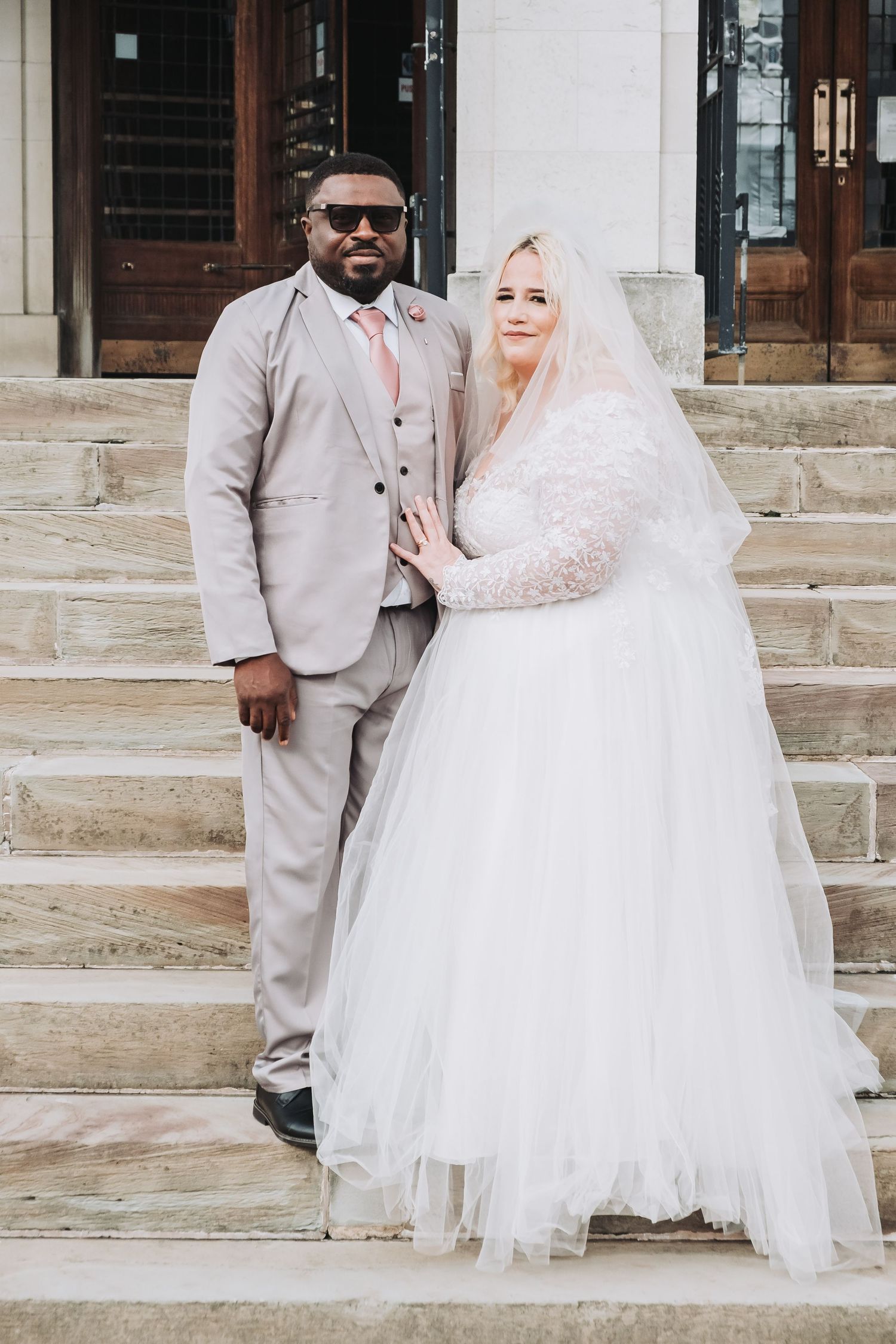 A newlywed couple in white dress and light gray suit pose on stone steps outside a building.