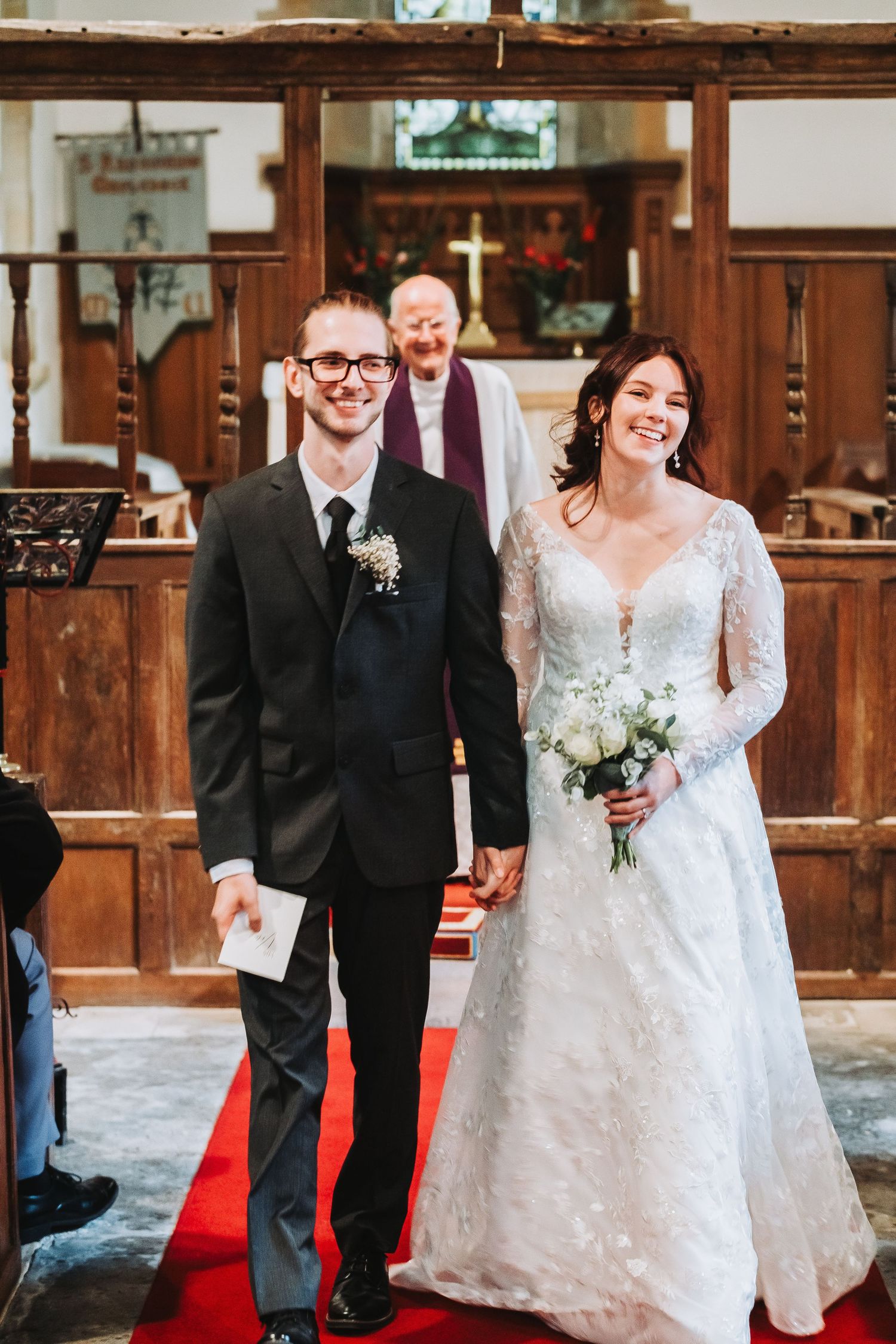 A wedding couple walks down a red carpet aisle in a rustic wooden church venue during their ceremony.