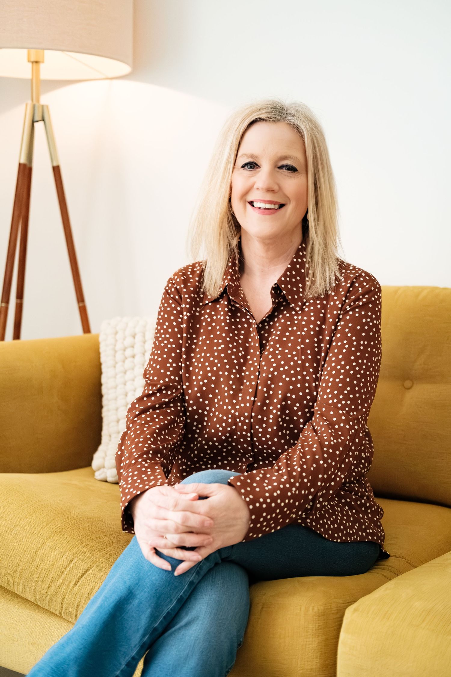 Smiling blonde woman in brown polka dot blouse sitting on yellow sofa holding a mug in a bright living room.