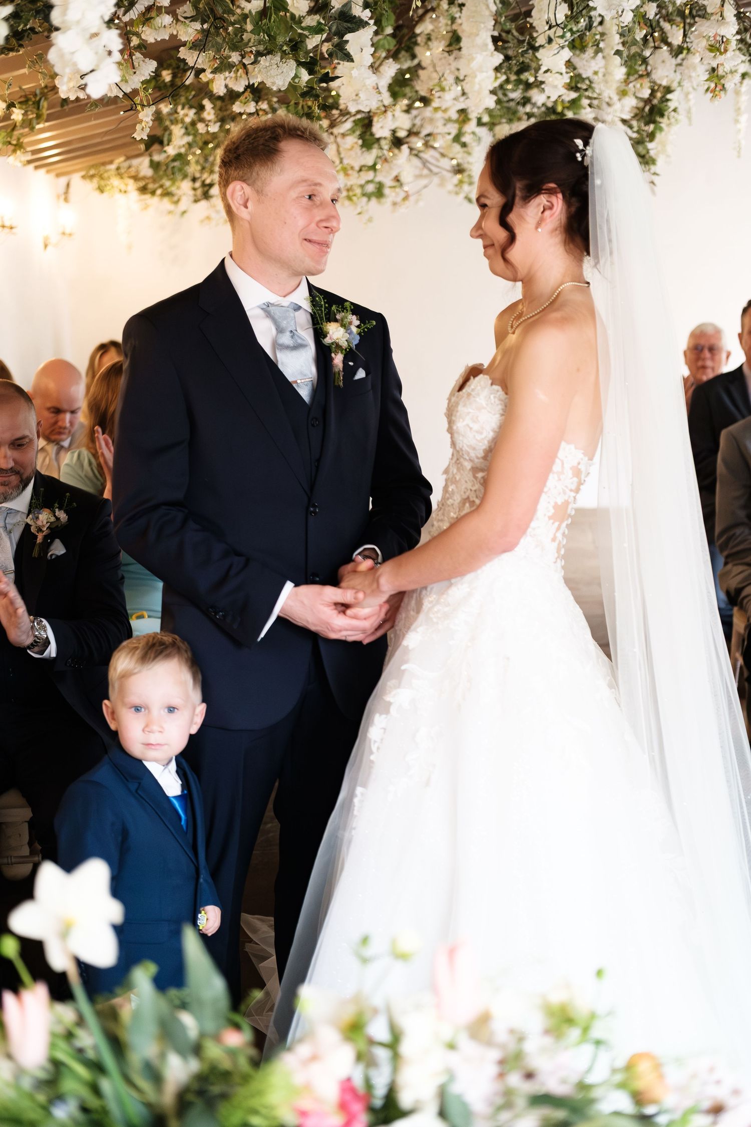Bride and groom exchange vows at floral wedding ceremony with young ring bearer in navy suit.