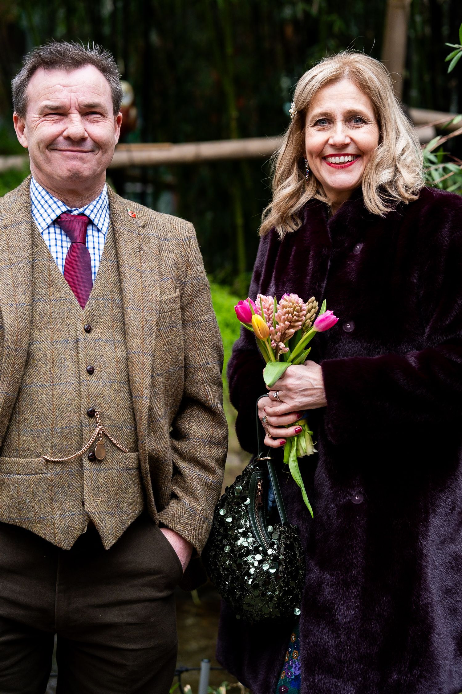 A couple in formal wear standing together outdoors with greenery in the background.