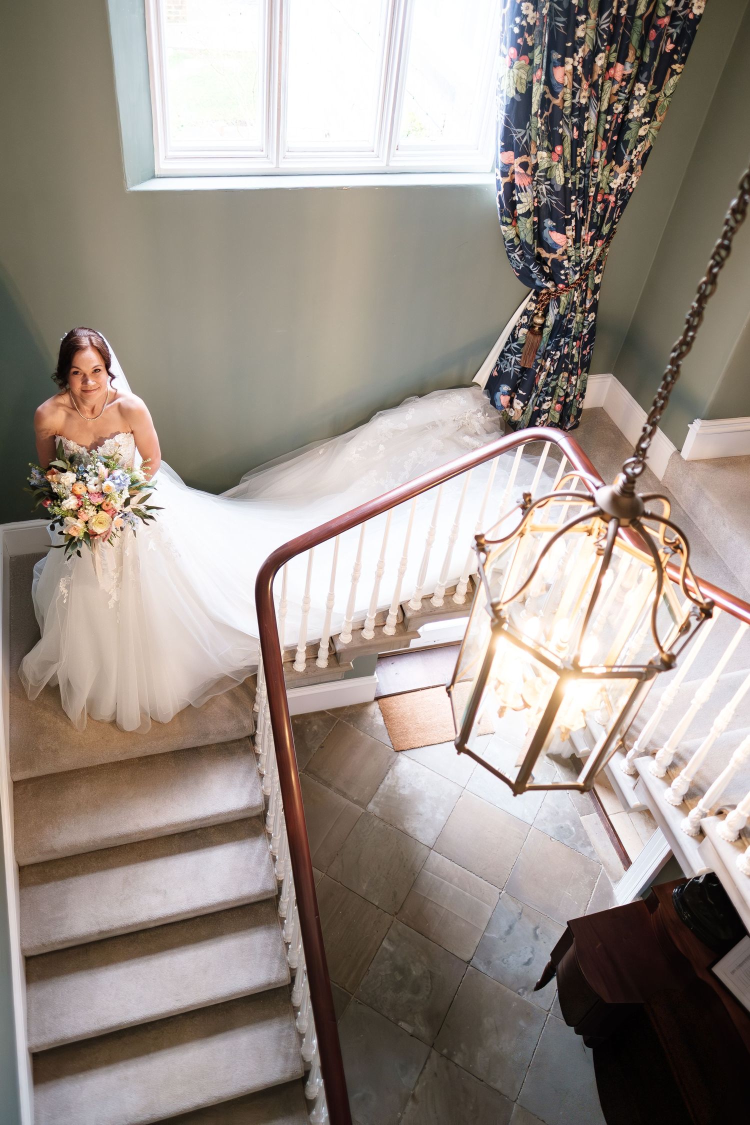 Bride in white ball gown holding bouquet descends elegant staircase with brass lantern chandelier in grand venue  at dunston hall estate